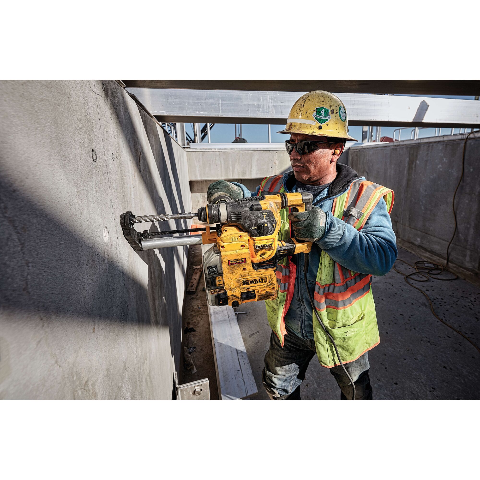 A construction worker wearing a yellow hard hat and reflective safety vest uses a DEWALT rotary hammer drill to drill into a concrete wall at a construction site.