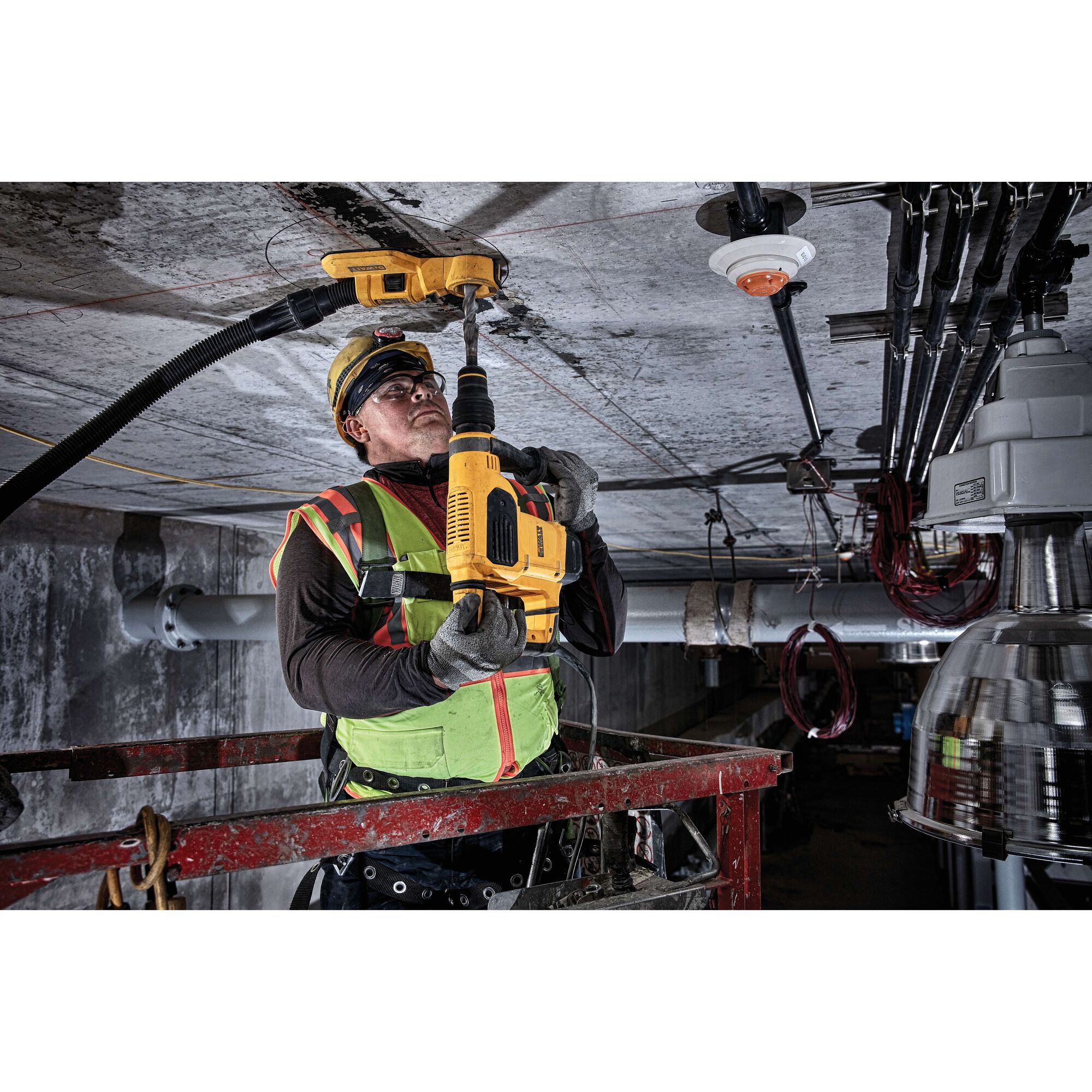 A worker in safety gear uses a yellow DEWALT rotary hammer drill (SKU: D25481K) to drill into a concrete ceiling in an industrial environment. The drill is connected to a dust extraction system.