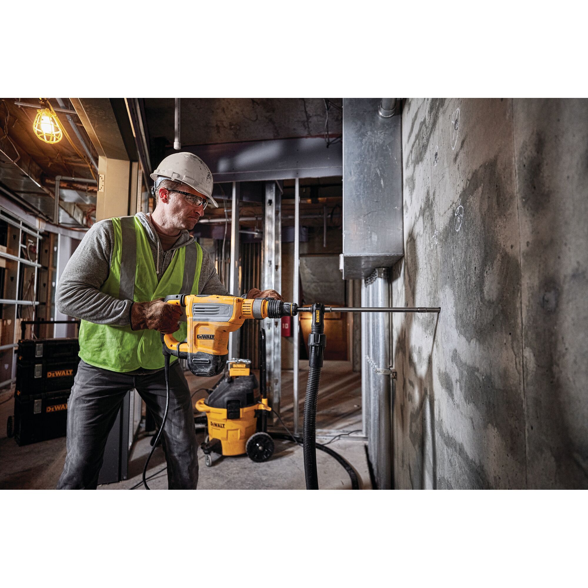 A worker wearing a safety helmet and vest operates a DEWALT rotary hammer drill (SKU: D25614K) to drill into a concrete wall at a construction site. DEWALT equipment and tools are visible in the background.