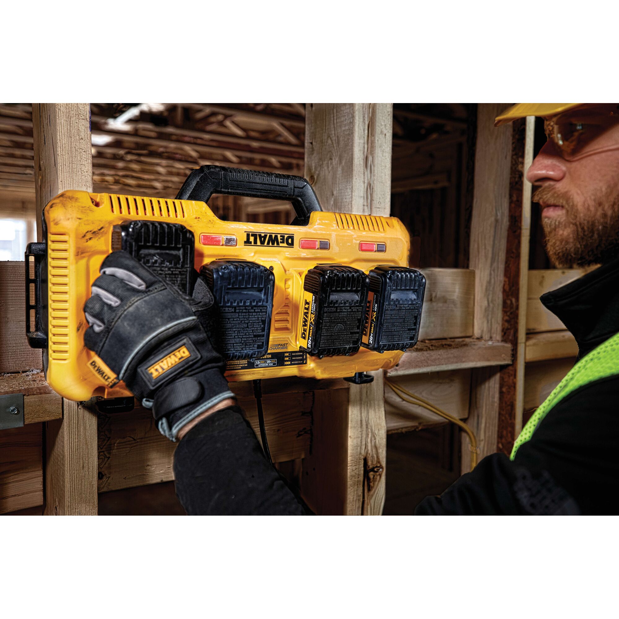 A Dewalt DCB104 yellow multiport battery charger mounted on a wooden beam at a construction site. A person wearing Dewalt-branded gloves is handling the charger, and there are four Dewalt batteries attached to it.