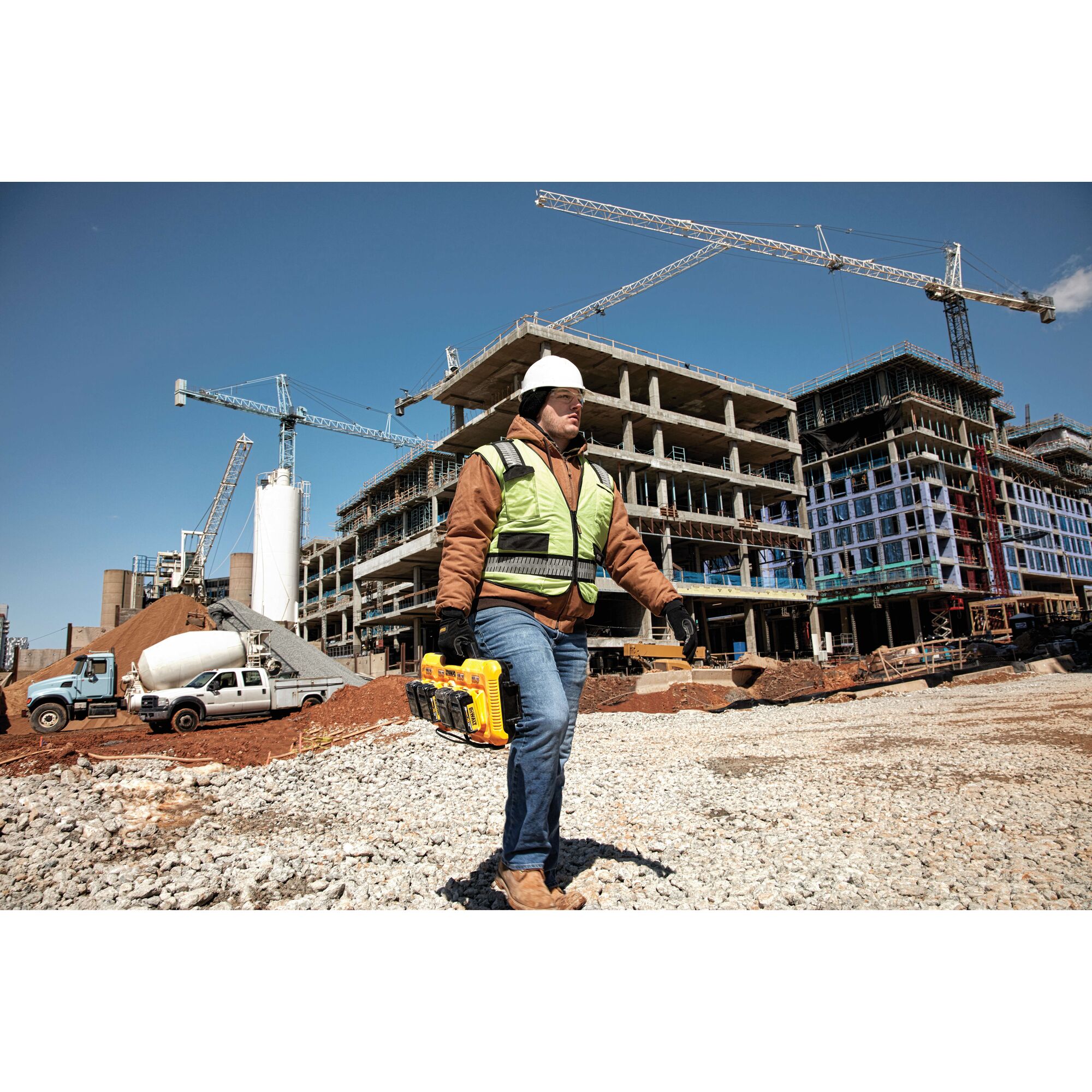 A construction worker wearing a safety vest and hard hat is carrying a yellow DEWALT DCB104 battery charger with batteries at a construction site. Several cranes and unfinished buildings are visible in the background.