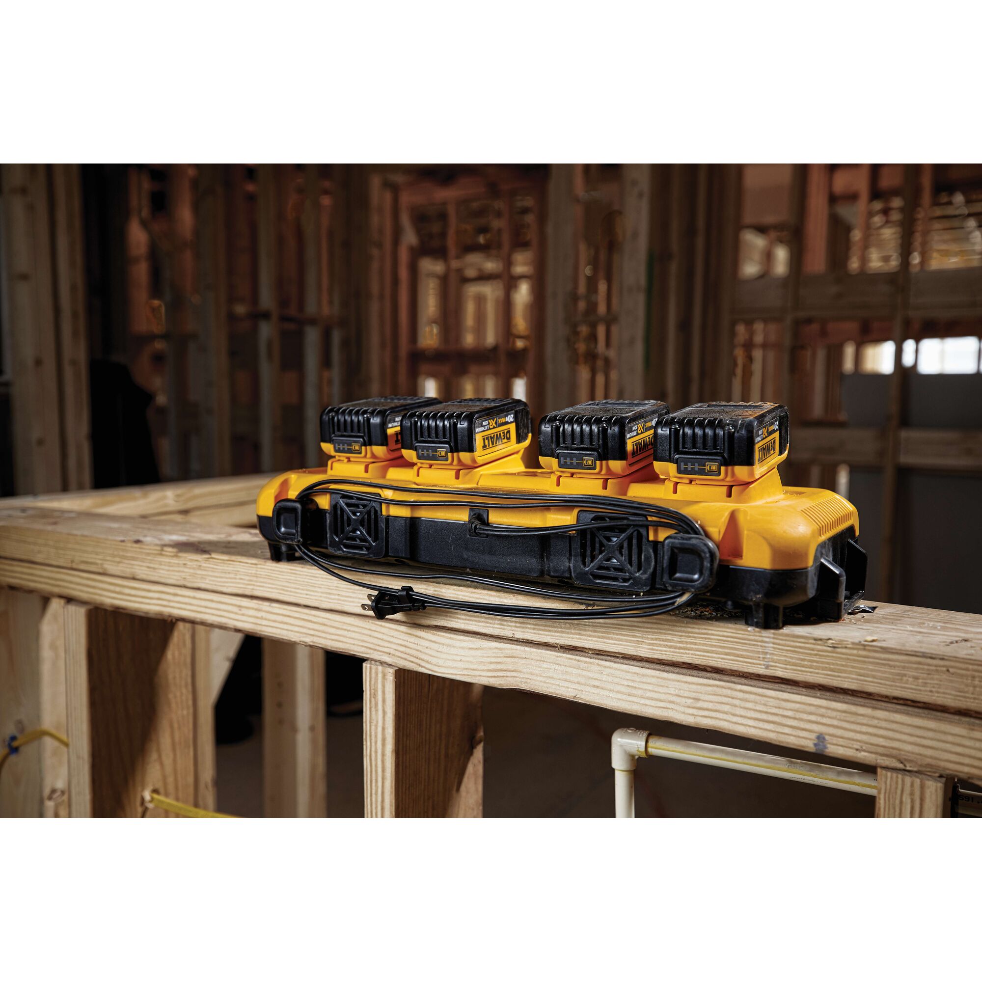 A DEWALT DCB104 yellow four-port battery charger with four DEWALT batteries, placed on a wooden beam in an unfinished construction site.