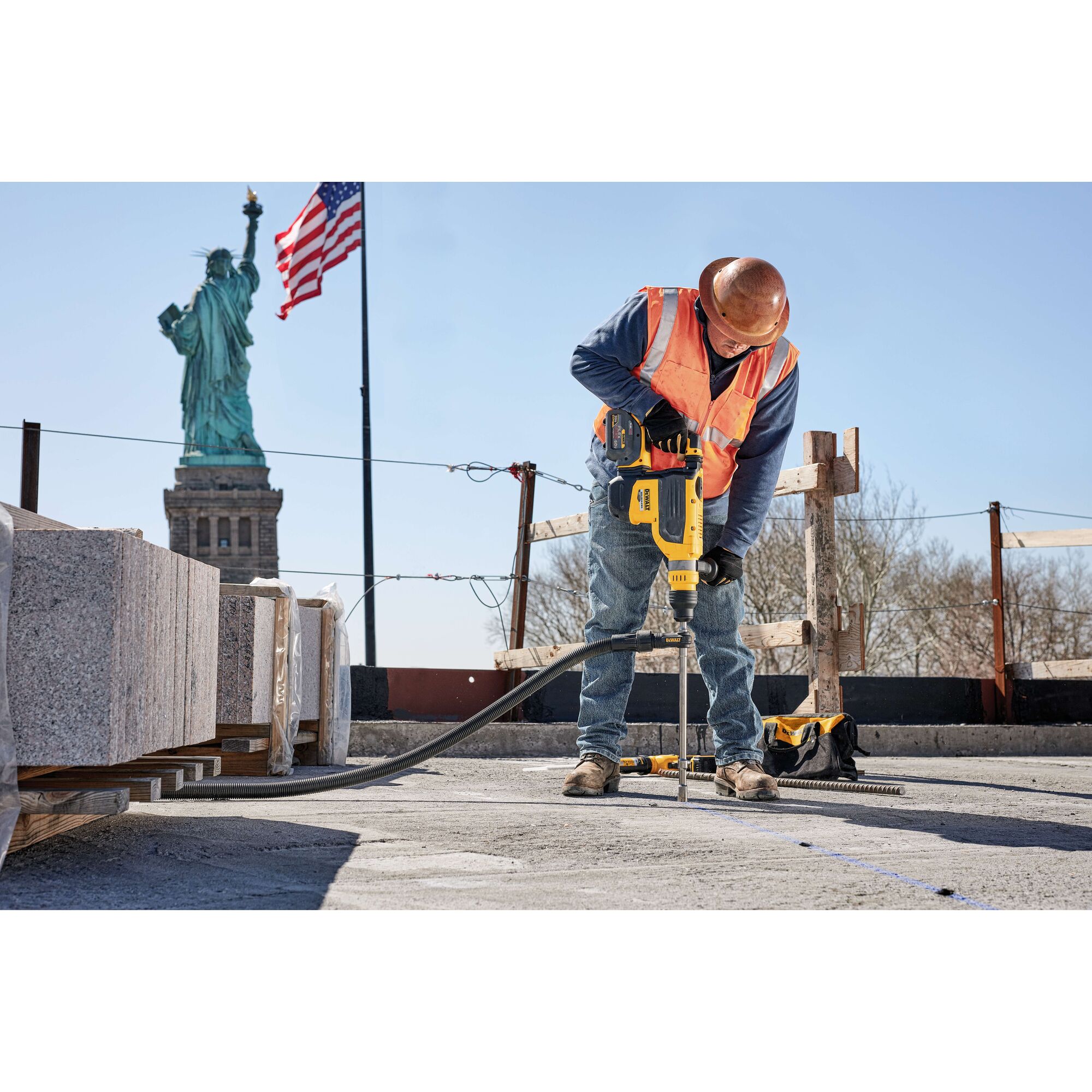 A construction worker wearing a hard hat and orange safety vest is using a yellow DEWALT power tool to drill into a concrete surface at a job site. Granite blocks and construction materials are visible nearby, with the Statue of Liberty and an American flag in the background.