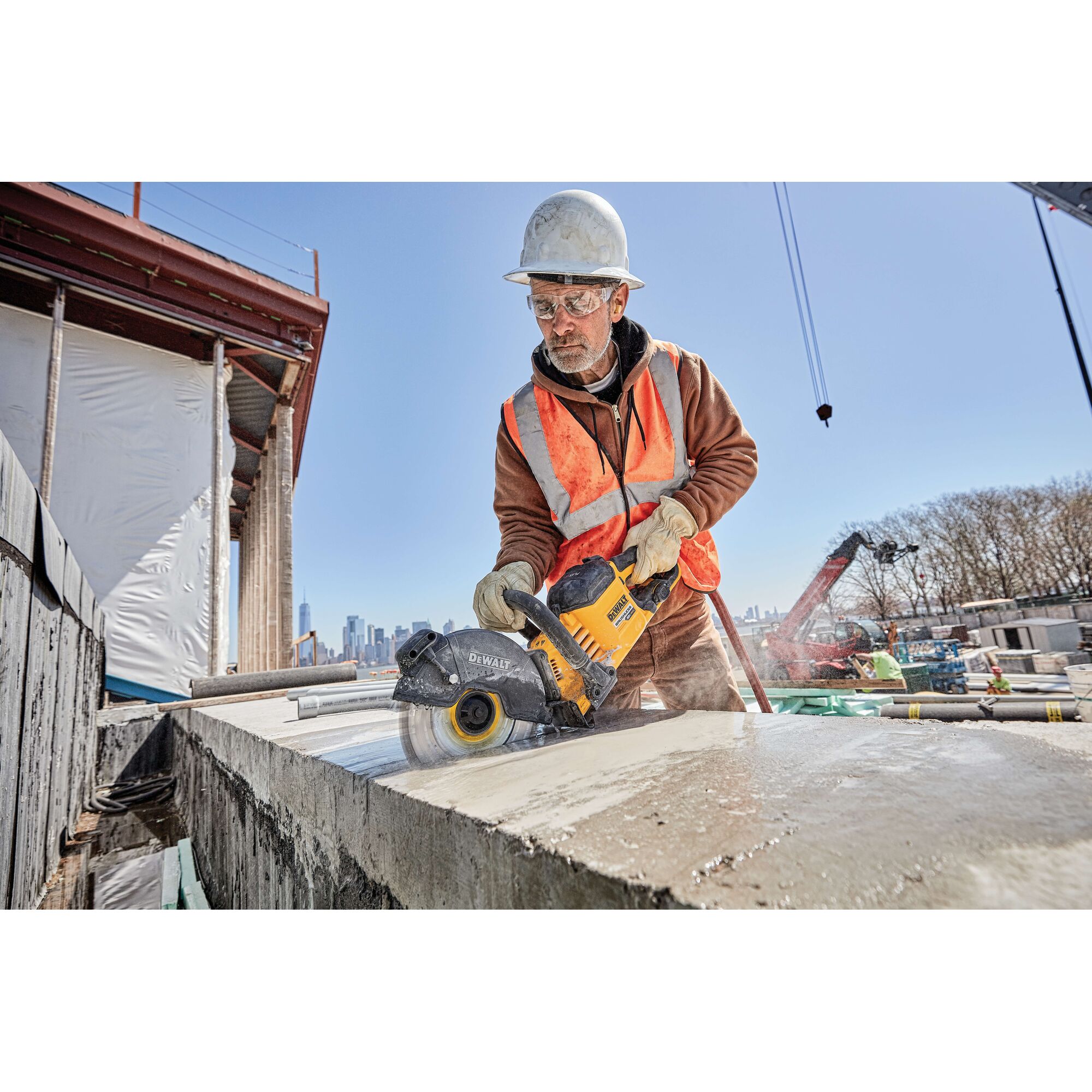 A construction worker wearing a safety vest, gloves, and a helmet is using a DEWALT power tool to cut concrete on a construction site. The worker’s face is blurred, and the DEWALT tool is clearly visible. The background shows ongoing construction and a clear sky.