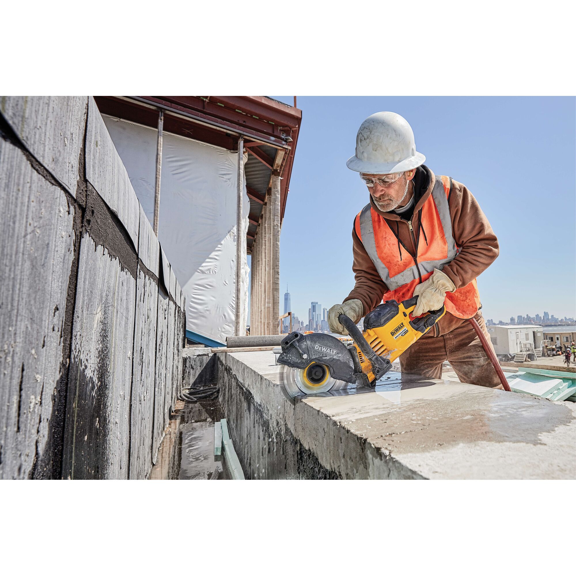 A construction worker wearing a hard hat and safety vest is using a yellow DEWALT DCB612 power tool to cut through a concrete block at an outdoor building site. The worker's face is blurred for privacy.