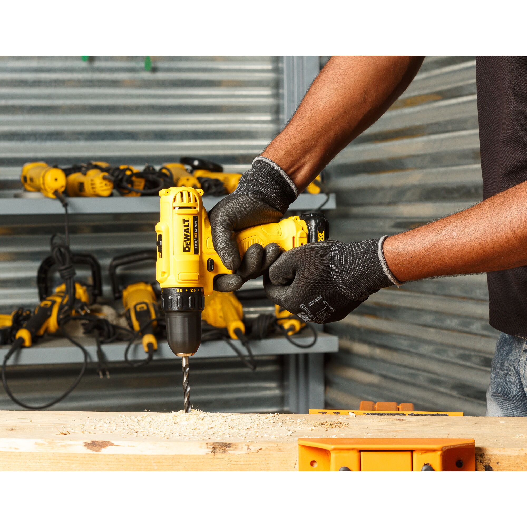 A person wearing black gloves uses a yellow DEWALT cordless drill to drill into a piece of wood. In the background, several power tools are visible on shelves in a workshop.