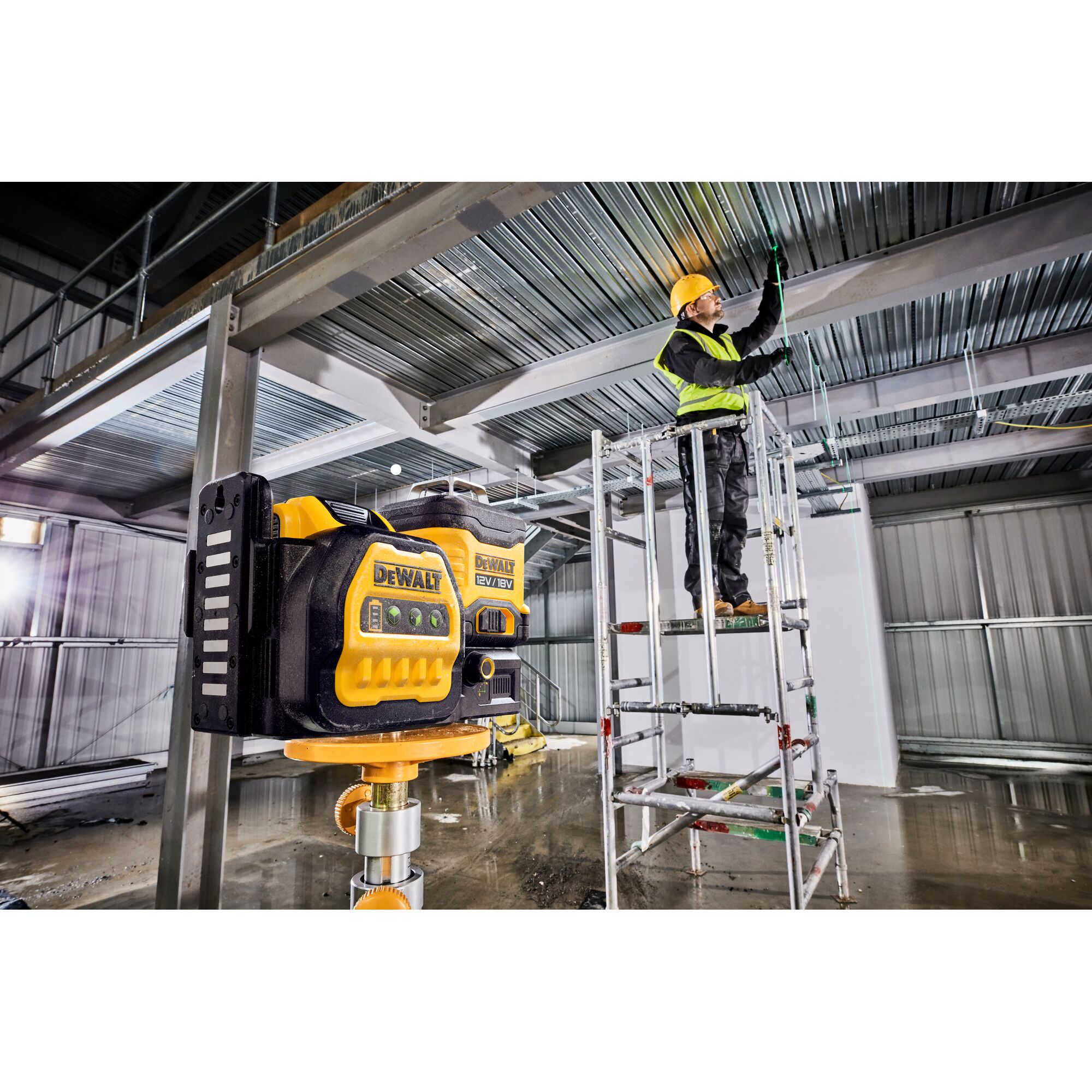 A DEWALT DCE089NG18 laser level is mounted on a tripod inside an industrial building under construction. In the background, a worker wearing a hard hat and safety vest stands on a scaffold, installing wiring along the metal ceiling beams.