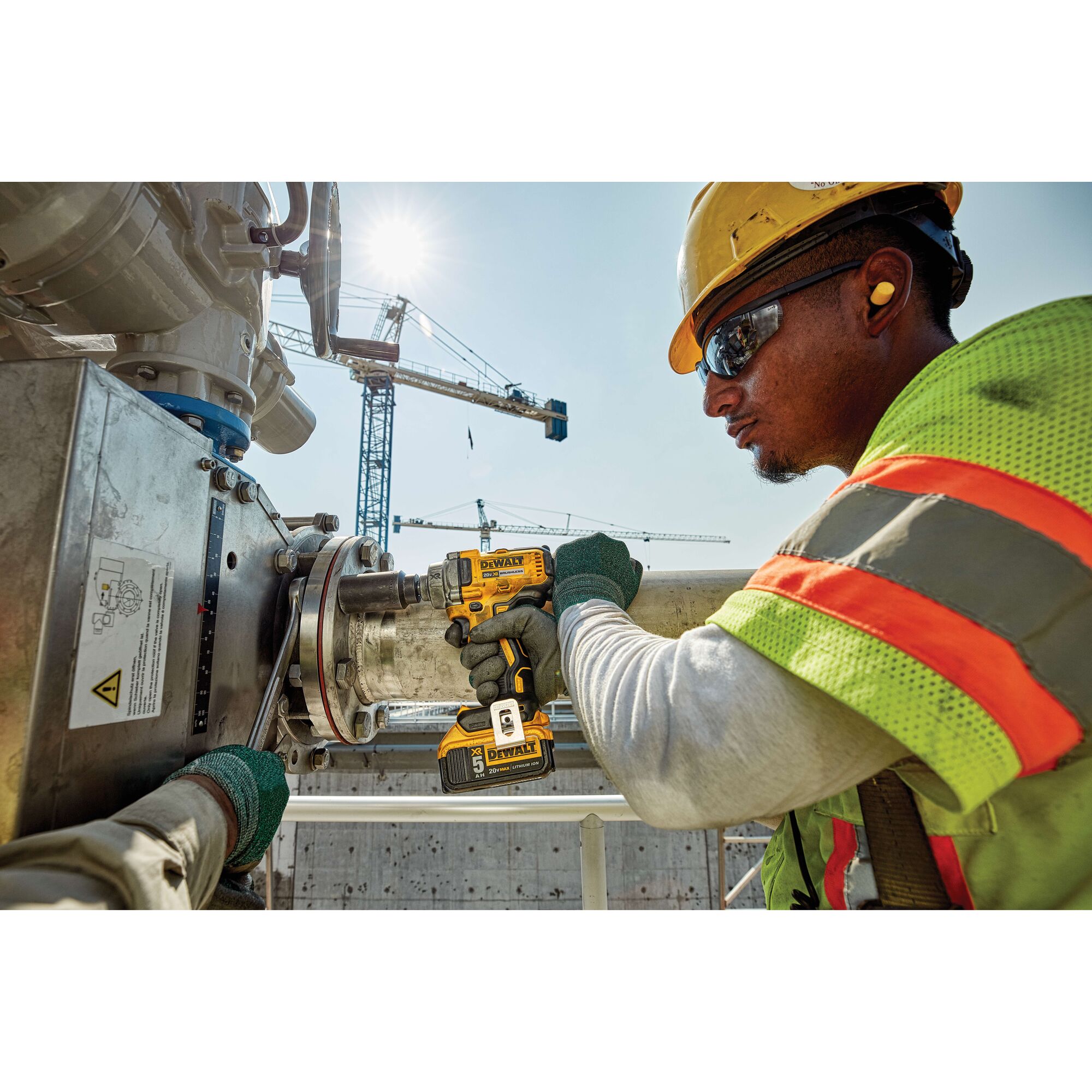 Worker uses DEWALT DCF894P2 impact wrench on metal pipe at construction site with cranes in background.