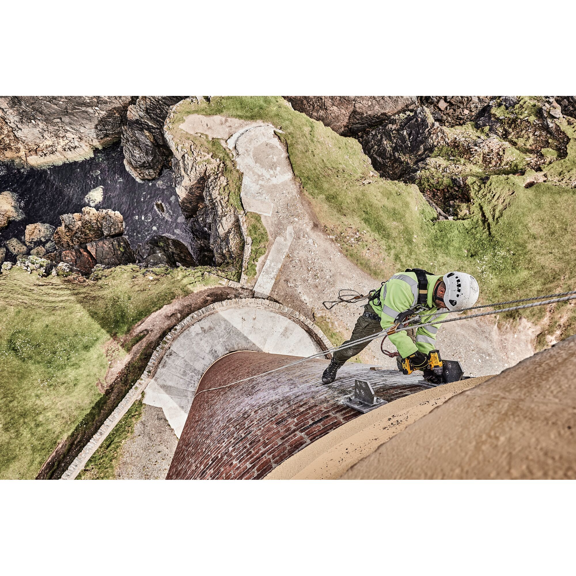 A worker wearing safety gear is using a DEWALT power tool while suspended from ropes on the side of a tall brick structure, with rocky terrain and water visible below.
