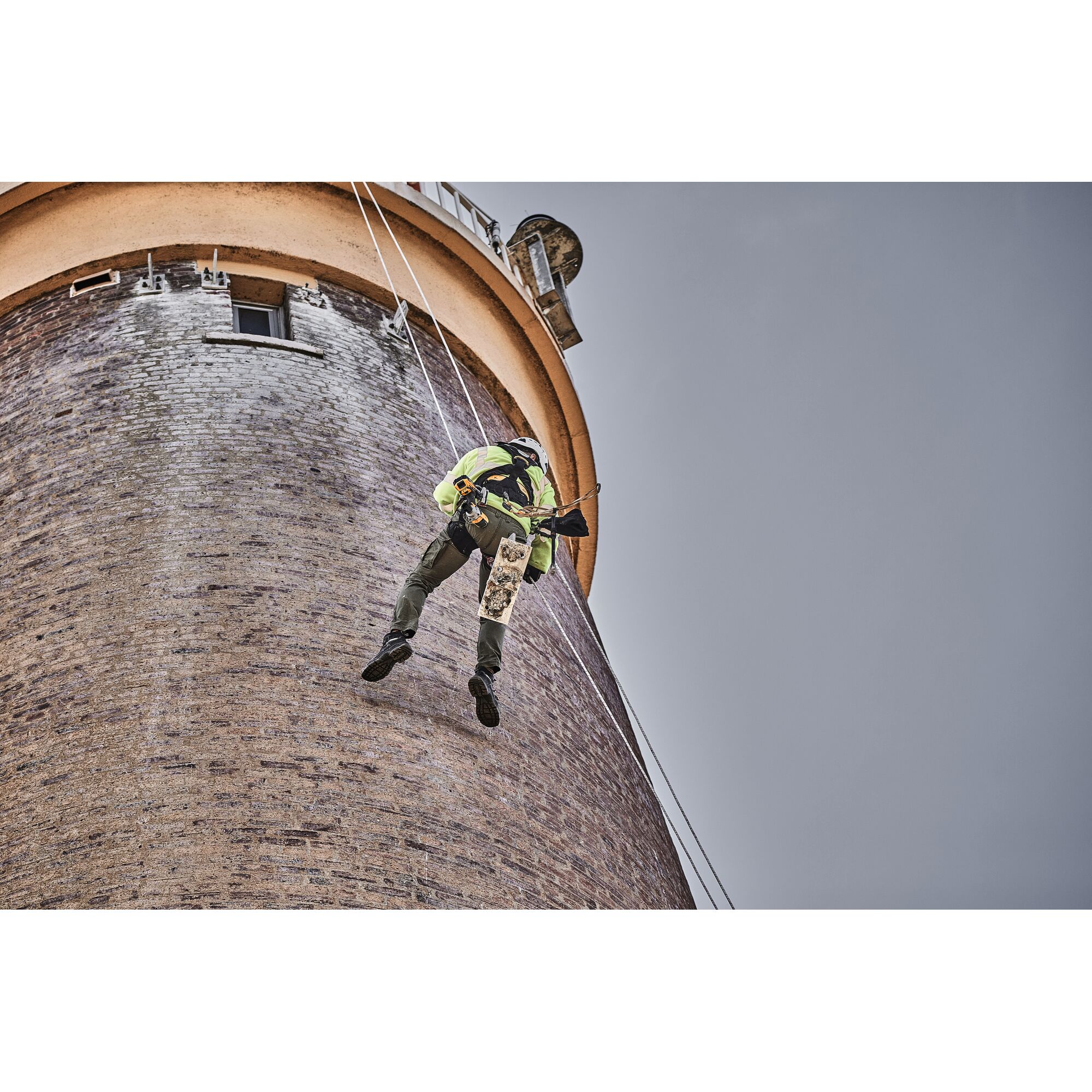 A worker wearing safety gear is hanging by ropes on the side of a tall brick building, using a DEWALT power tool.