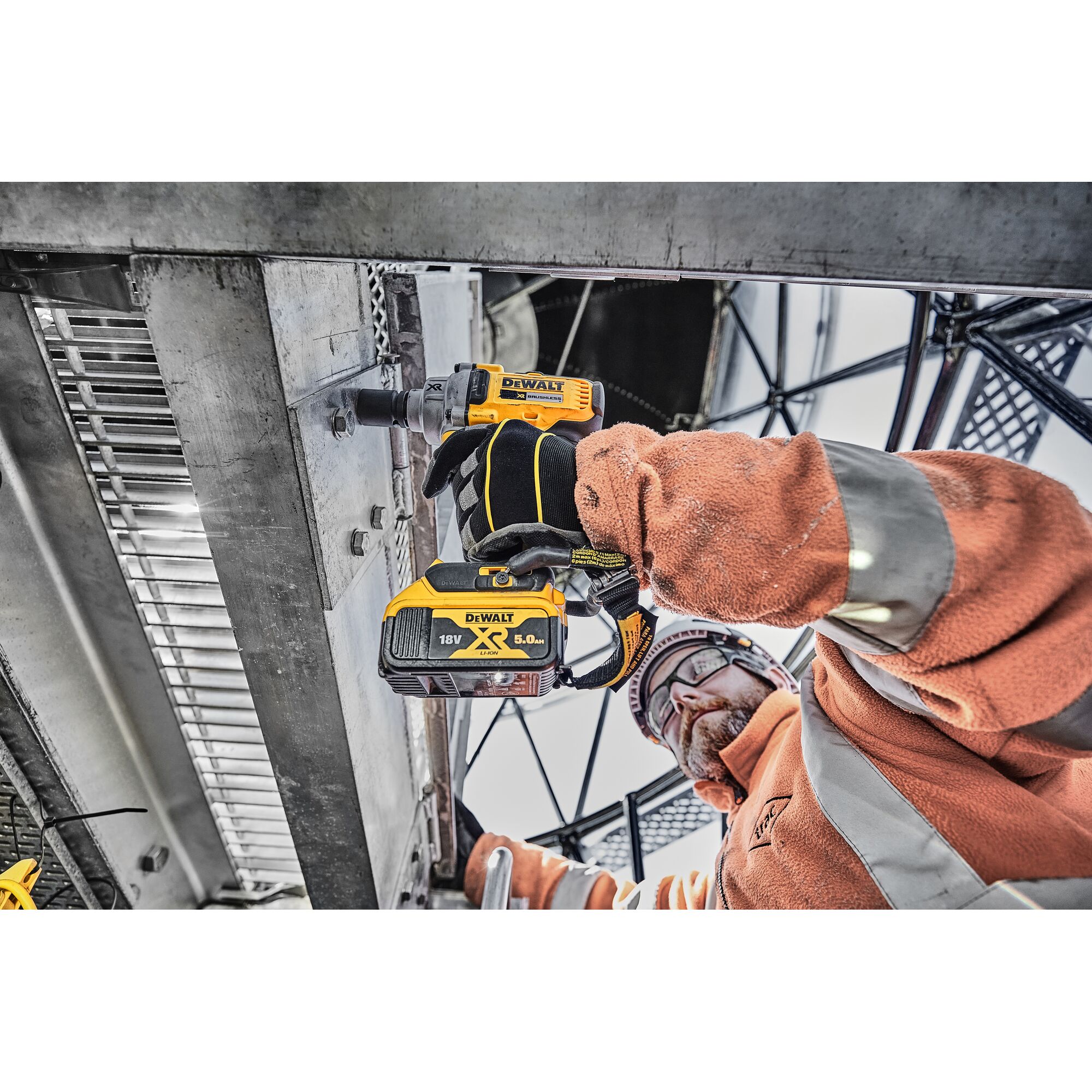 A worker wearing an orange safety jacket and gloves uses a DEWALT DCF894P2 cordless impact wrench to fasten bolts on a metal structure at an industrial worksite.