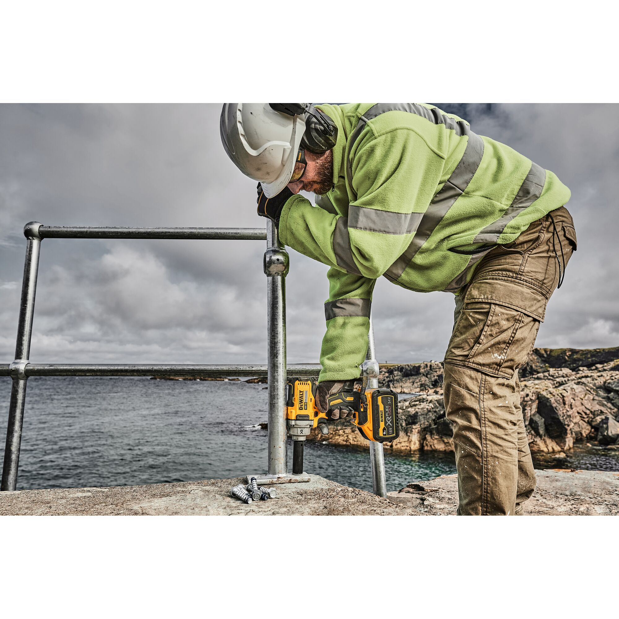 A person wearing safety gear, including a hard hat, high-visibility jacket, and safety glasses, uses a DEWALT cordless impact wrench (SKU: DCF894P2) to fasten a metal railing outdoors near rocky terrain and water.