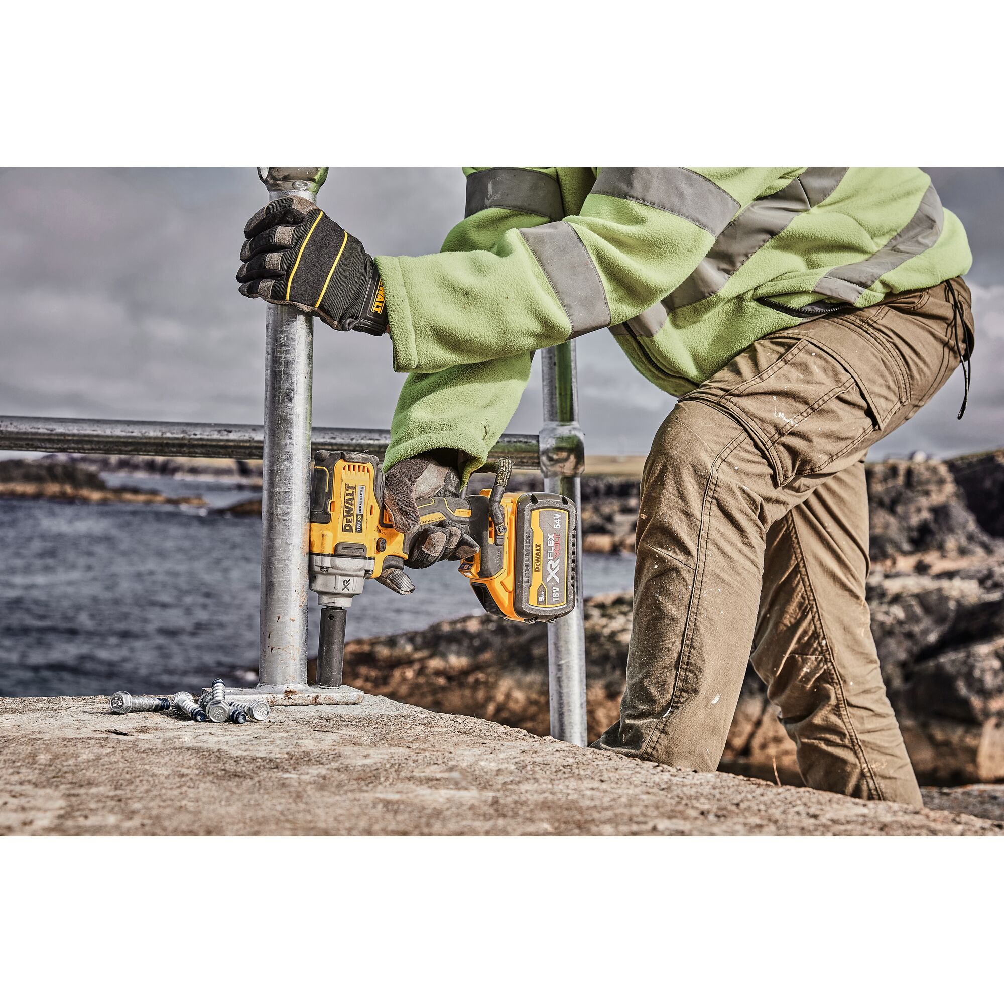 A person wearing work gloves and a high-visibility jacket is using a DEWALT cordless impact wrench (model DCF894P2) to secure a metal railing outdoors near a waterfront. Several bolts are lying on the concrete surface.