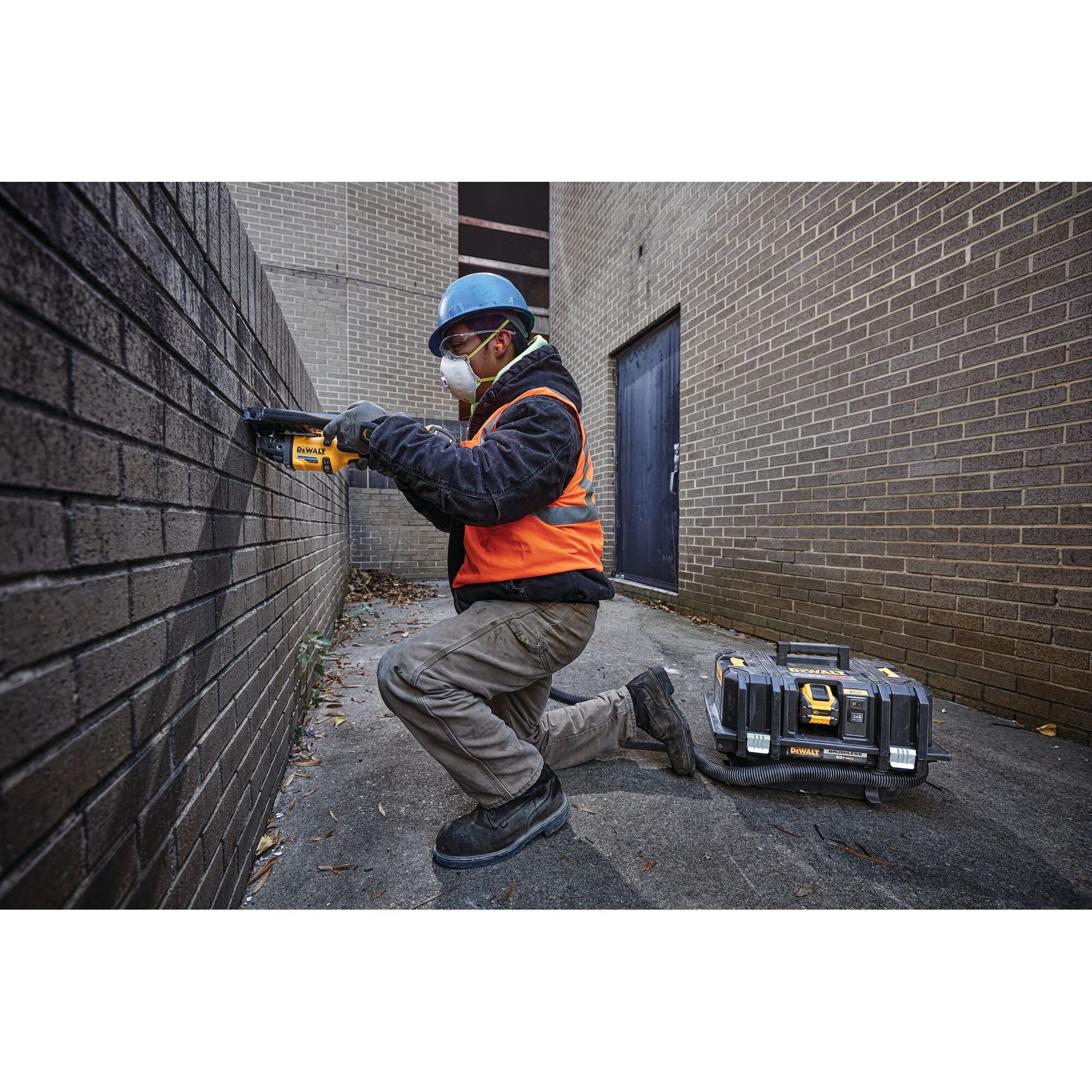 A construction worker wearing a blue safety helmet, orange vest, and gloves is kneeling outdoors using a DEWALT power tool on a brick wall. A DEWALT tool case is placed on the ground nearby.