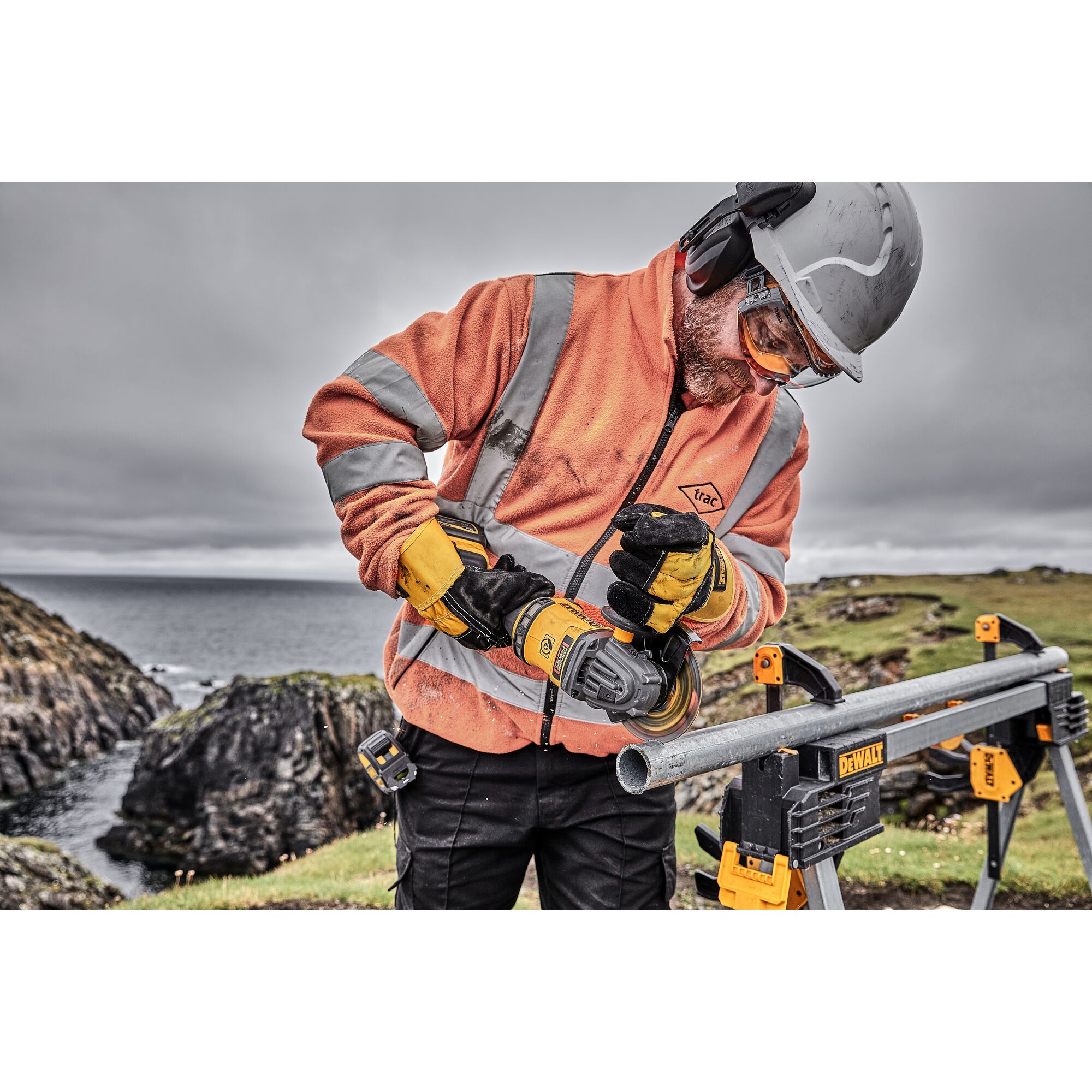 A worker wearing a safety helmet, goggles, earmuffs, and orange high-visibility jacket uses a DEWALT cordless angle grinder to cut a metal pipe outdoors. The pipe is secured on a DEWALT work stand, with a rocky coastline visible in the background.