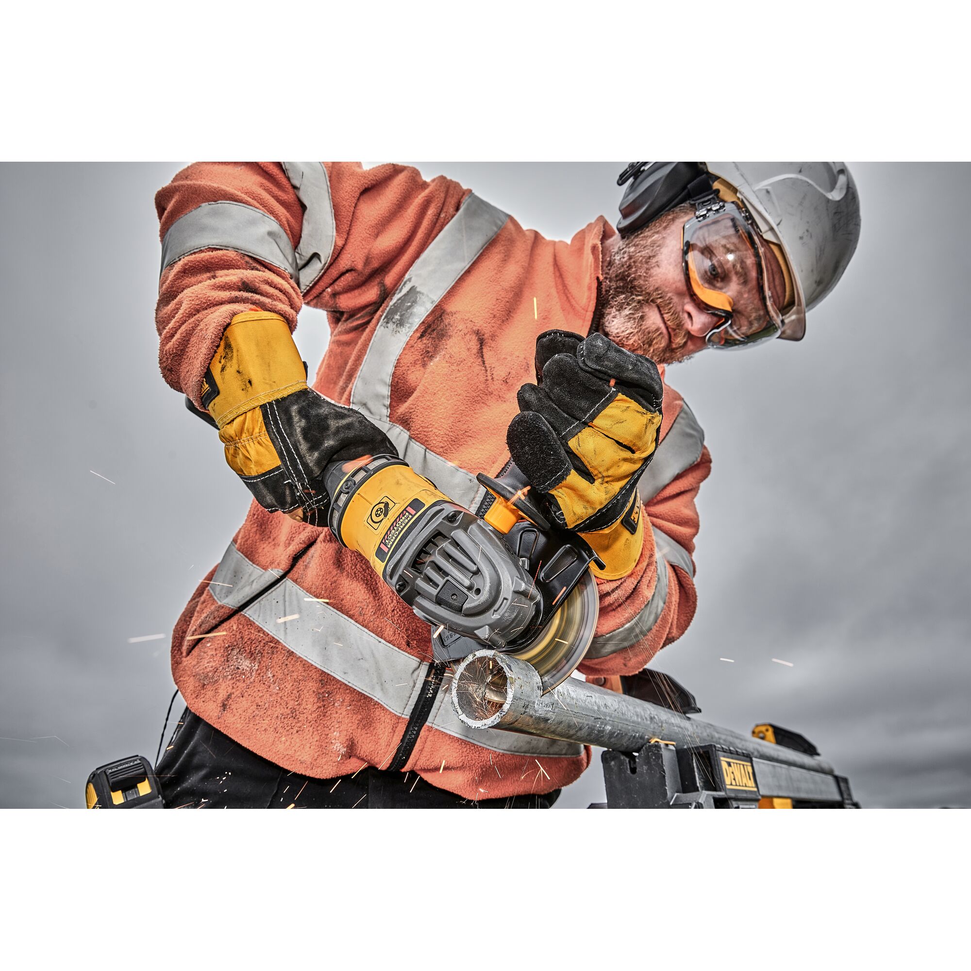 A construction worker wearing an orange high-visibility jacket, safety gloves, and a helmet is using a DEWALT cordless angle grinder to cut through a metal pipe, with visible sparks flying.