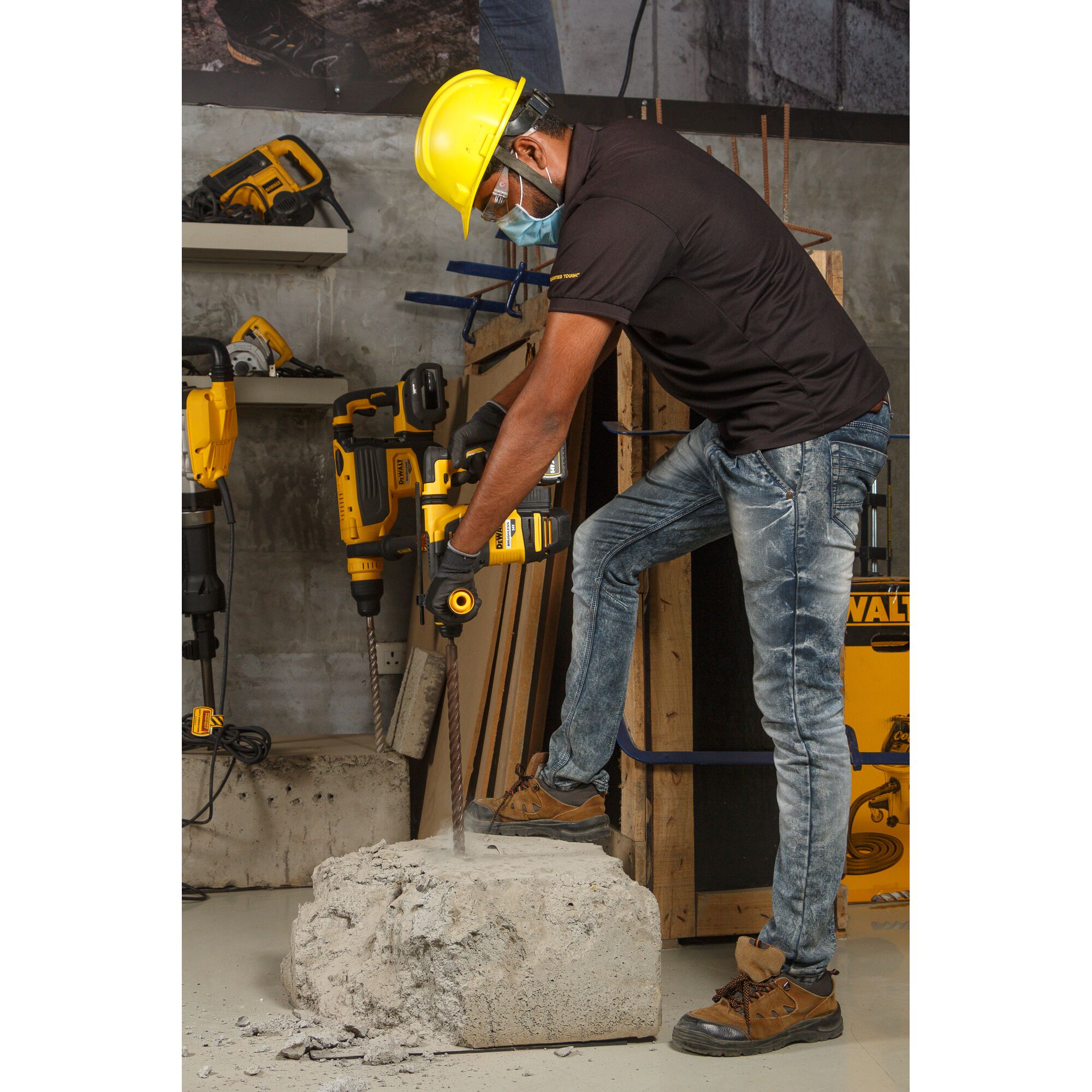 A person wearing a yellow safety helmet, gloves, and work boots is using a DEWALT DCH333 rotary hammer drill to work on a large concrete block indoors. The setting includes additional construction tools and materials visible in the background.