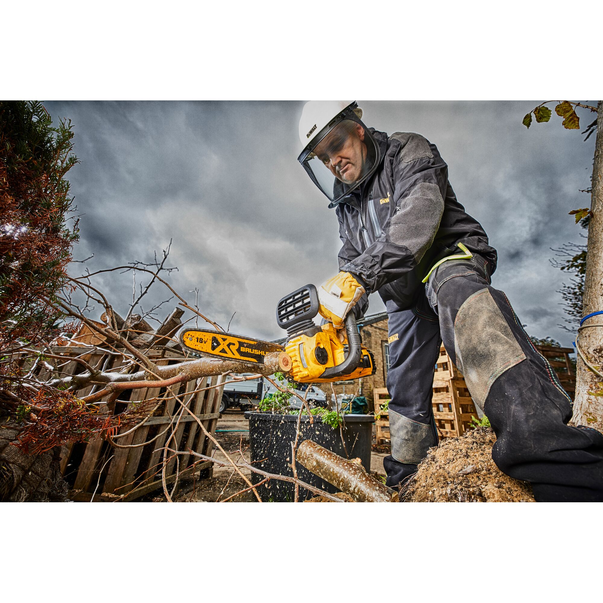 A person wearing safety gear uses a DEWALT DCM565N cordless brushless chainsaw to cut tree branches outdoors. The scene features cloudy skies, wooden pallets, and protective equipment.