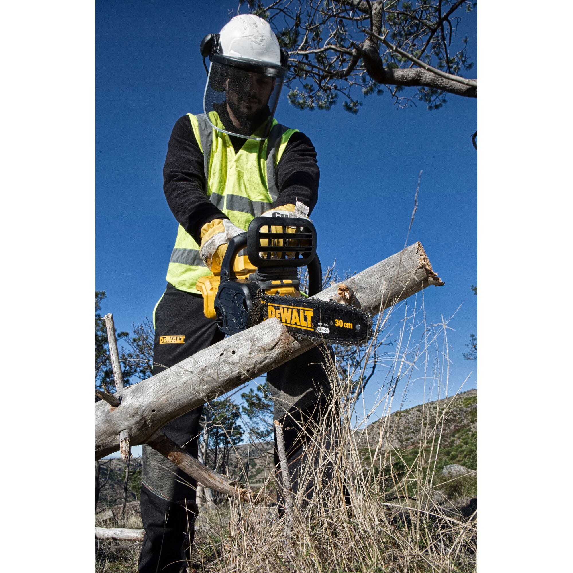 A person wearing protective gear and high-visibility clothing is using a DEWALT DCM565N chainsaw to cut a tree branch outdoors. The chainsaw has a visible 30 cm blade and is held securely, with the surroundings showing grass and trees.