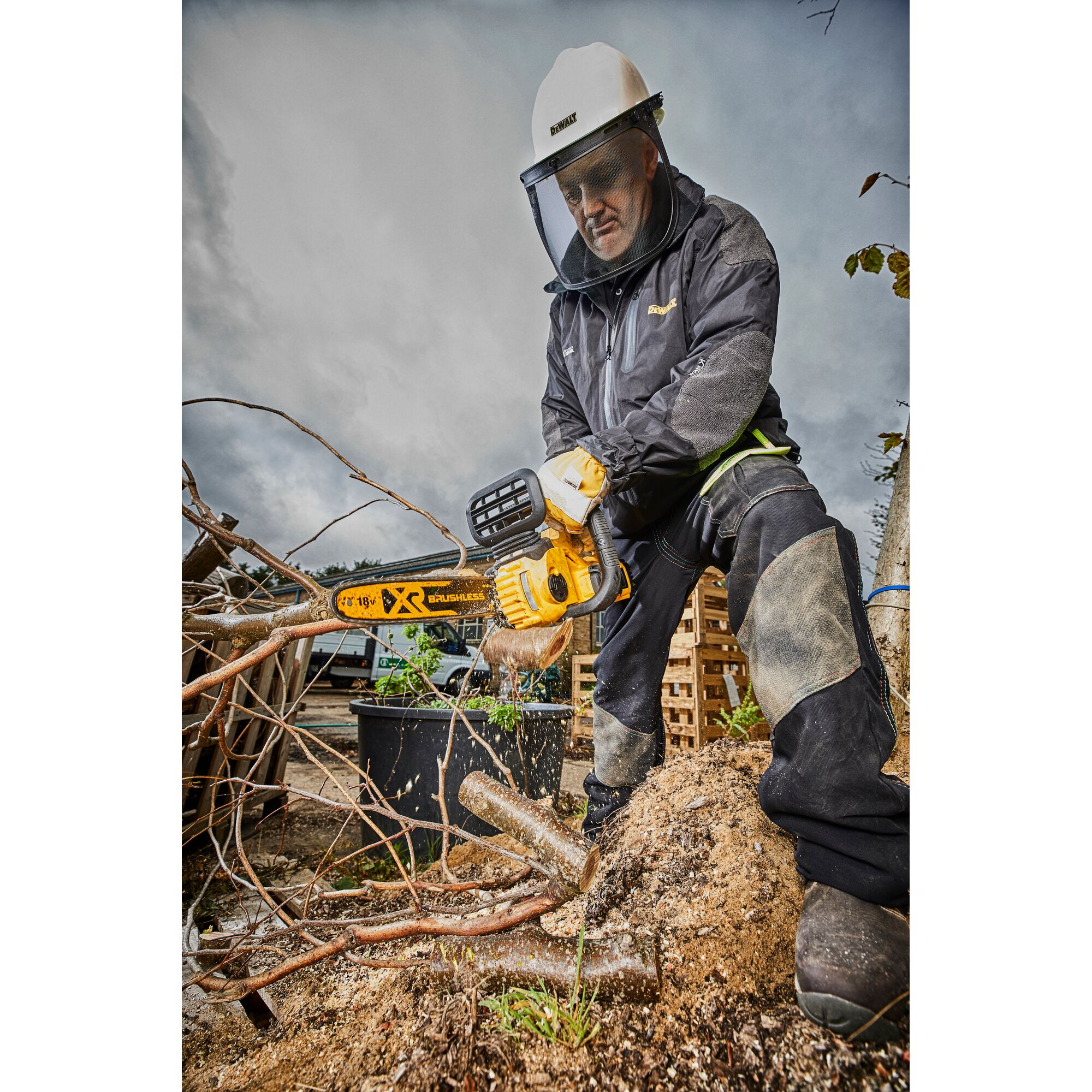 A person wearing protective gear uses a DEWALT DCM565N cordless chainsaw to cut tree branches outdoors. Sawdust is flying, and cut wood pieces are on the ground.