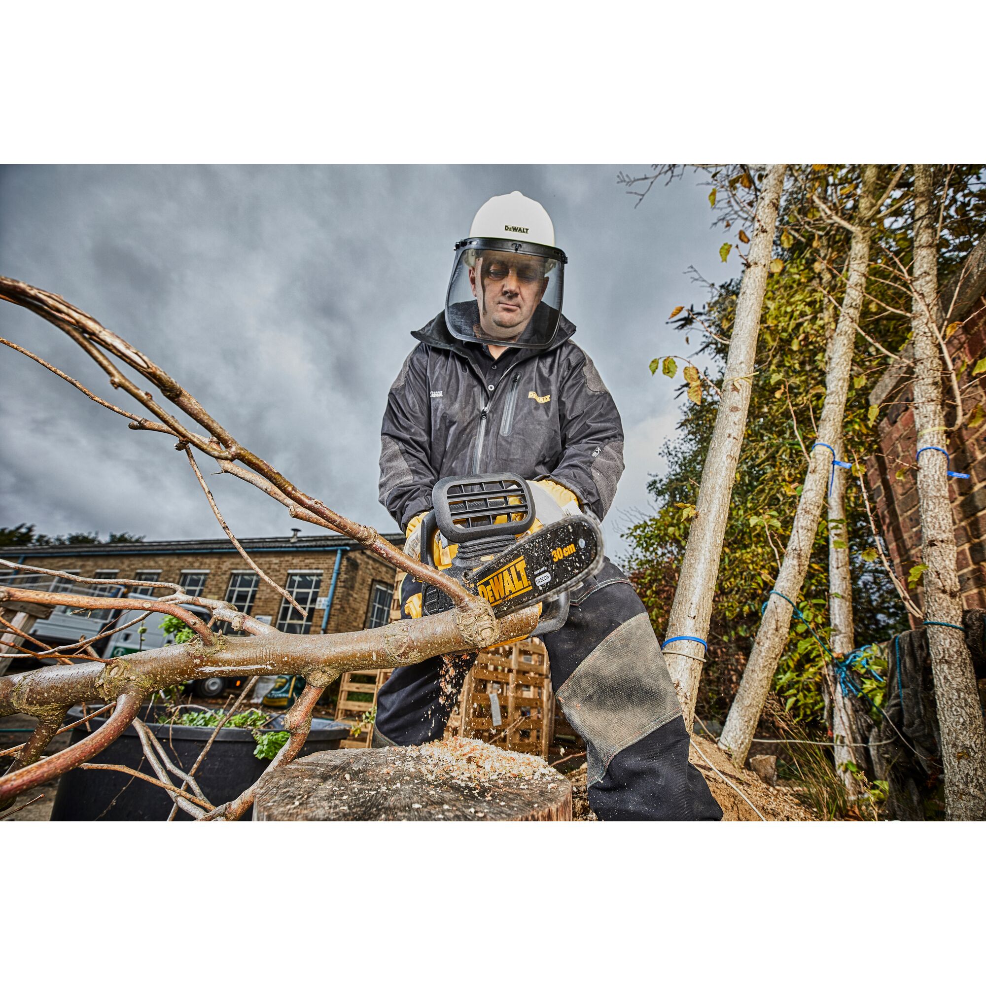 A person wearing protective gear, including a helmet and face shield, is using a DEWALT DCM565N cordless chainsaw to cut a tree branch outdoors. Sawdust is visible as the branch is being cut.