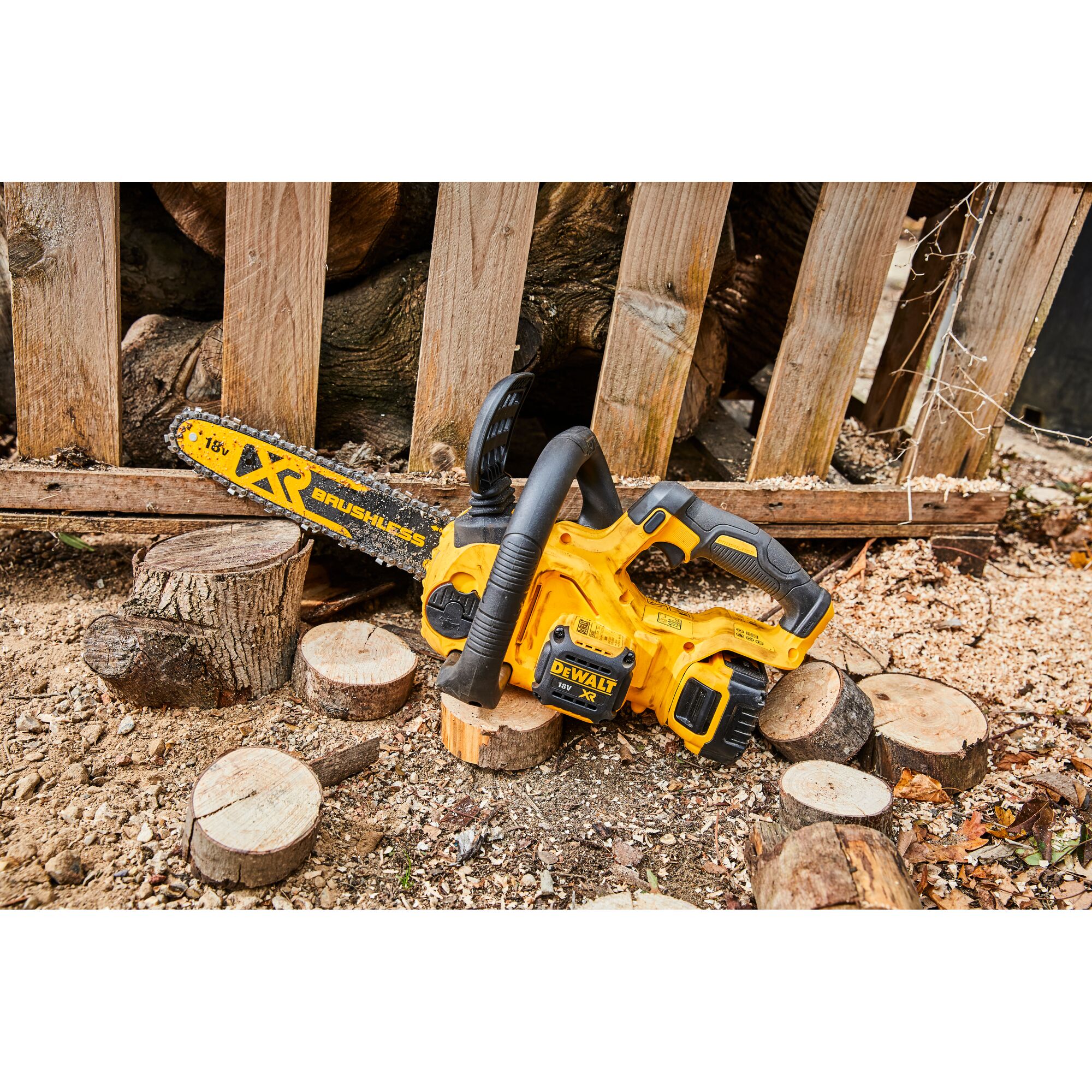 A yellow DEWALT XR brushless cordless chainsaw is placed on the ground surrounded by cut wooden logs and wood shavings. The chainsaw is fitted with an 18V battery and is resting outdoors in front of a wooden fence.