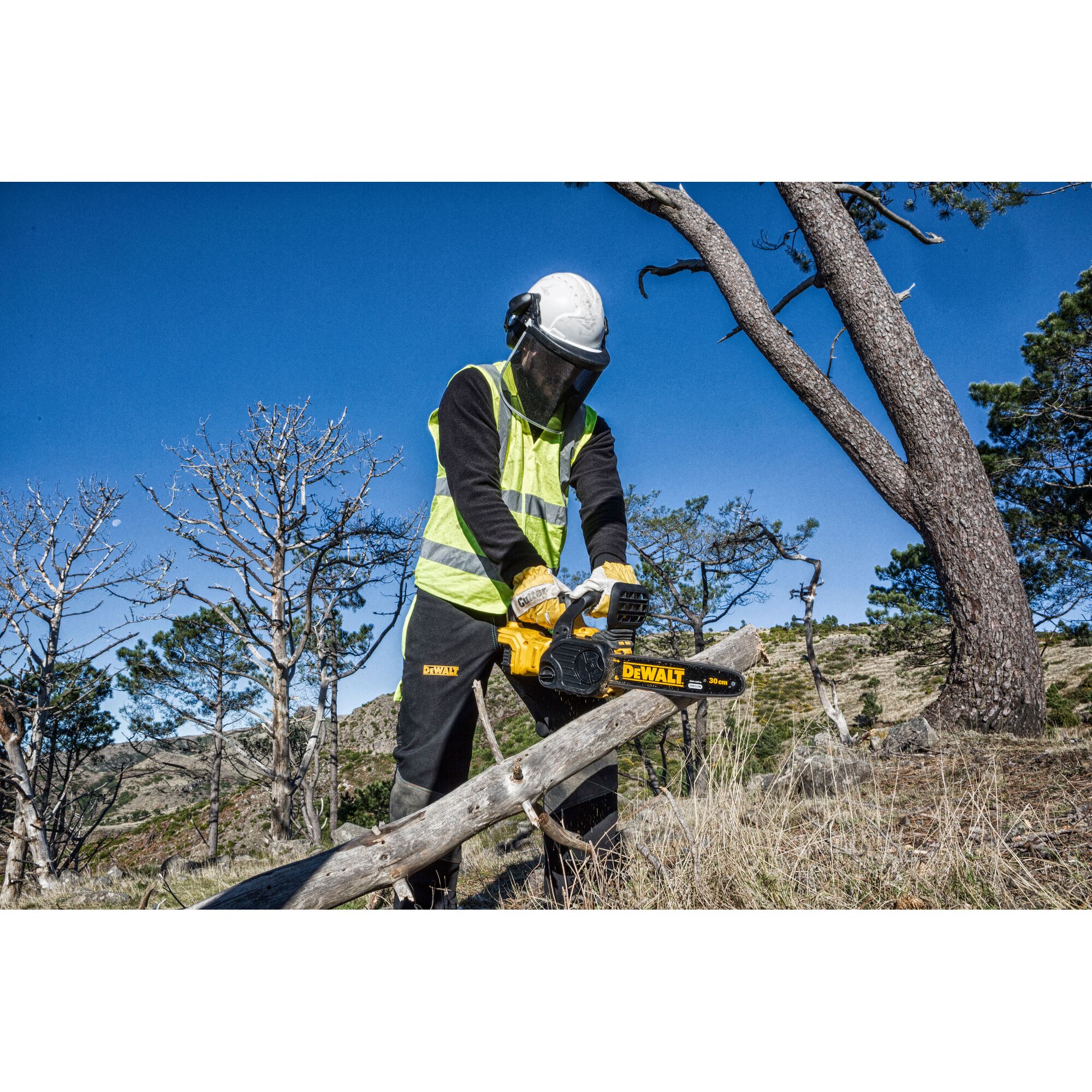 A person wearing safety gear and a high-visibility vest is using a DEWALT DCM565N chainsaw to cut a fallen tree branch outdoors in a forested area.