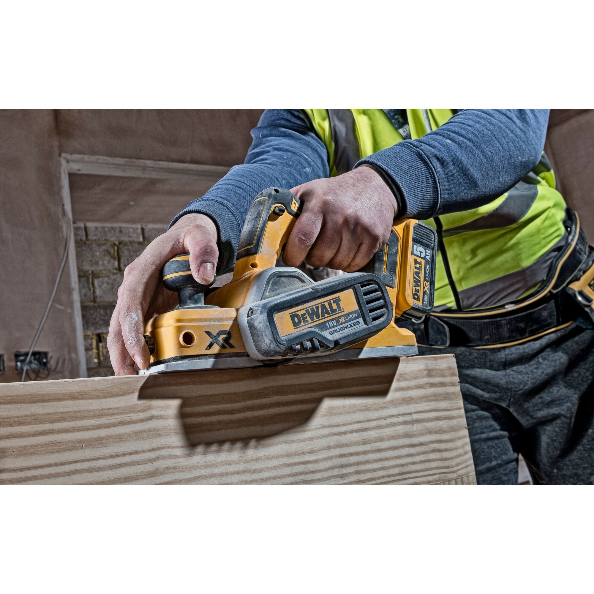 A person using a DEWALT DCP580N cordless planer to smooth a wooden board, wearing work clothes and safety gear in a workshop setting.