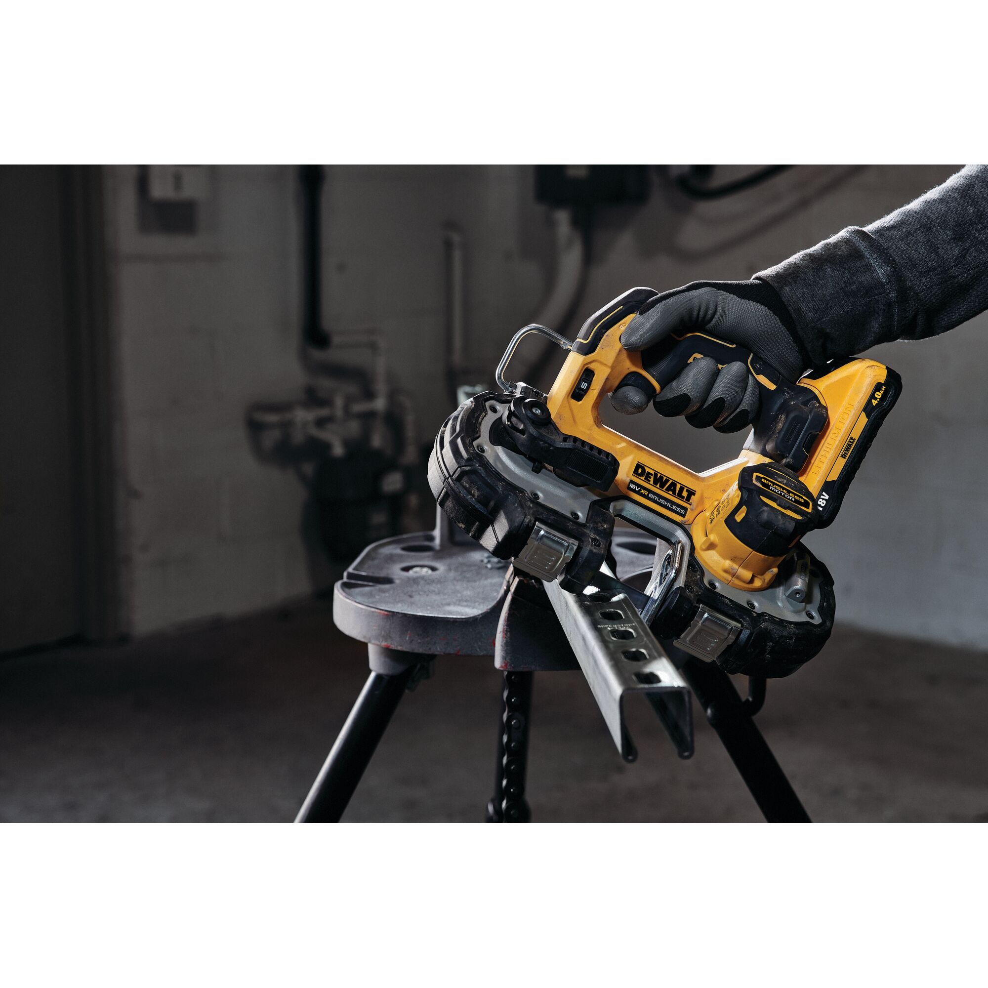 A person wearing gloves is using a yellow DEWALT cordless portable band saw to cut a metal piece that is resting on a workbench in an industrial setting.
