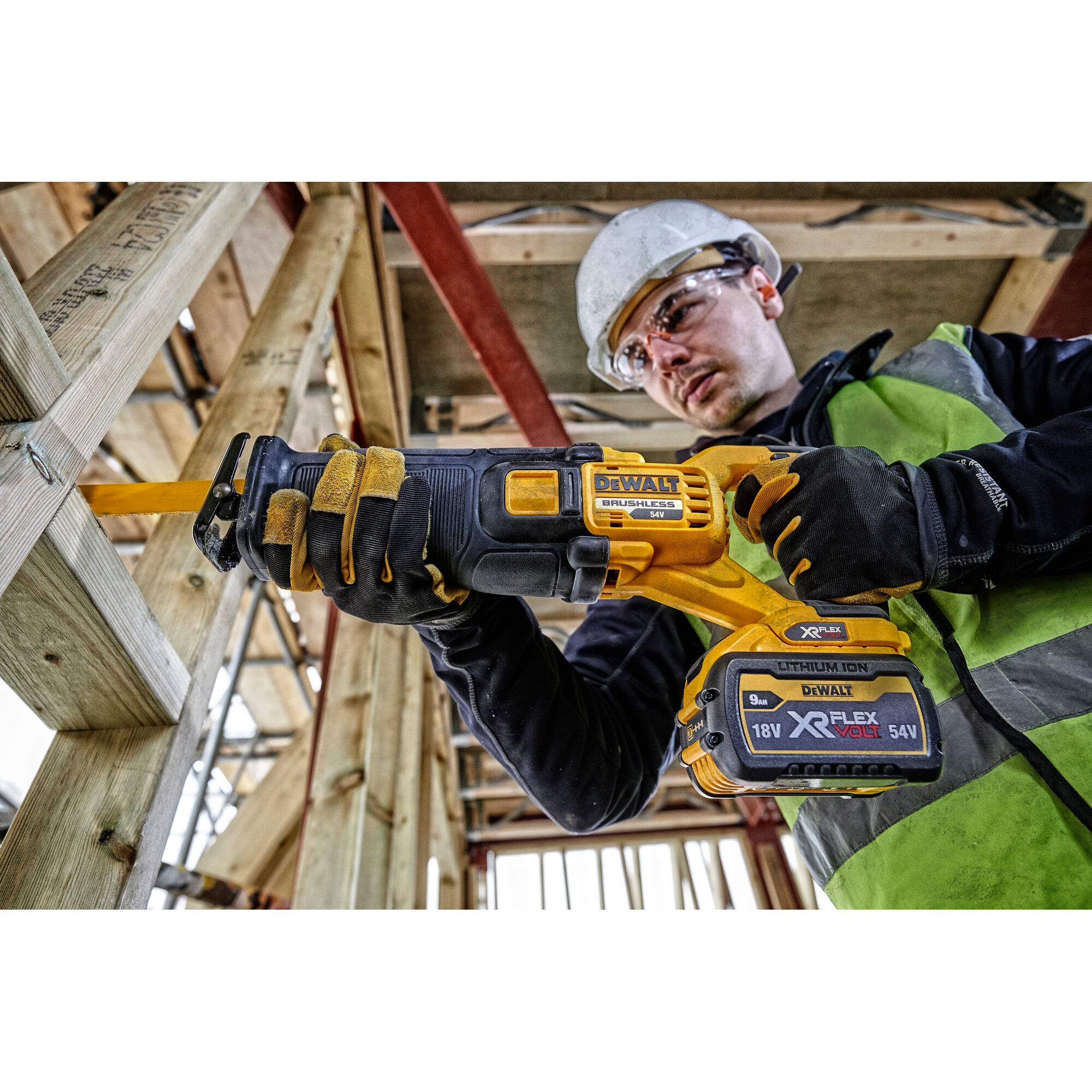 A person wearing safety gear uses a DEWALT cordless reciprocating saw, model DCS389N, to cut through wooden beams at a construction site. The tool is powered by an 18V/54V XR FLEXVOLT lithium-ion battery.