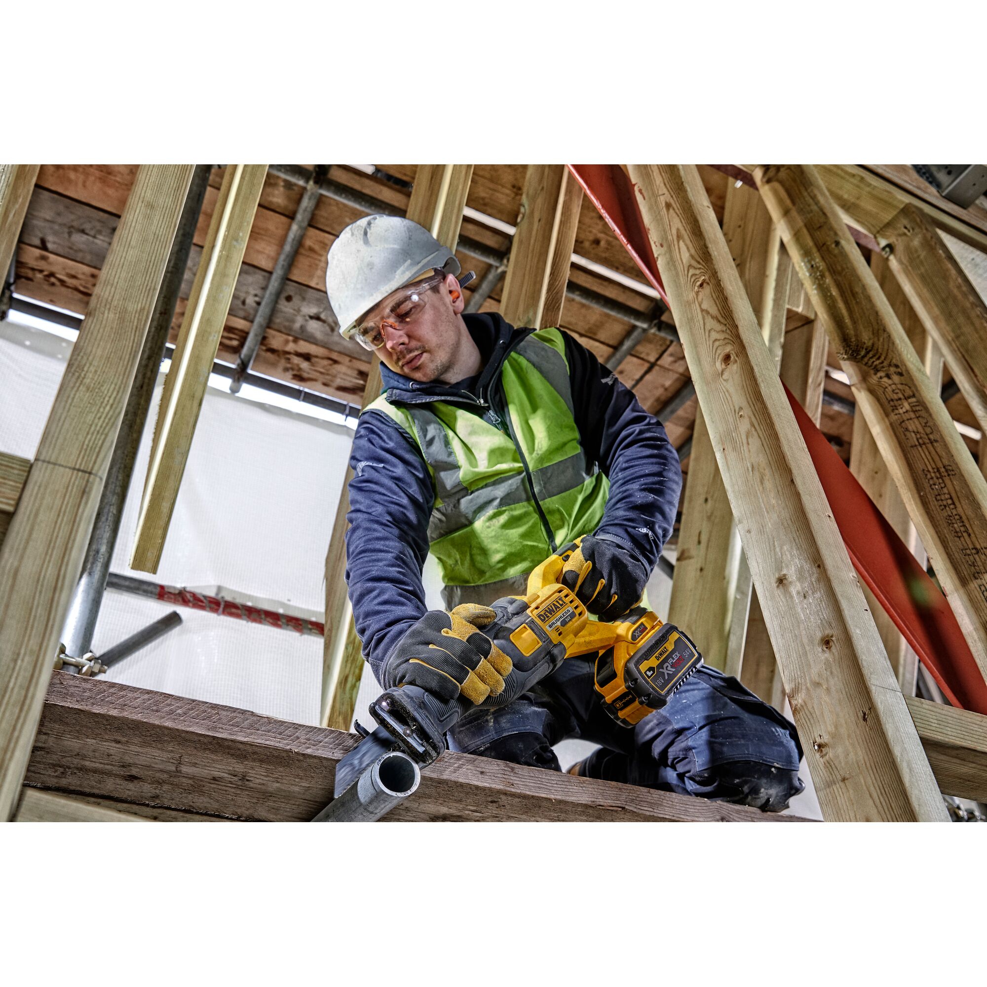 Person using a DEWALT cordless saw to cut pipe at a construction site, wearing safety gear and high-visibility vest.