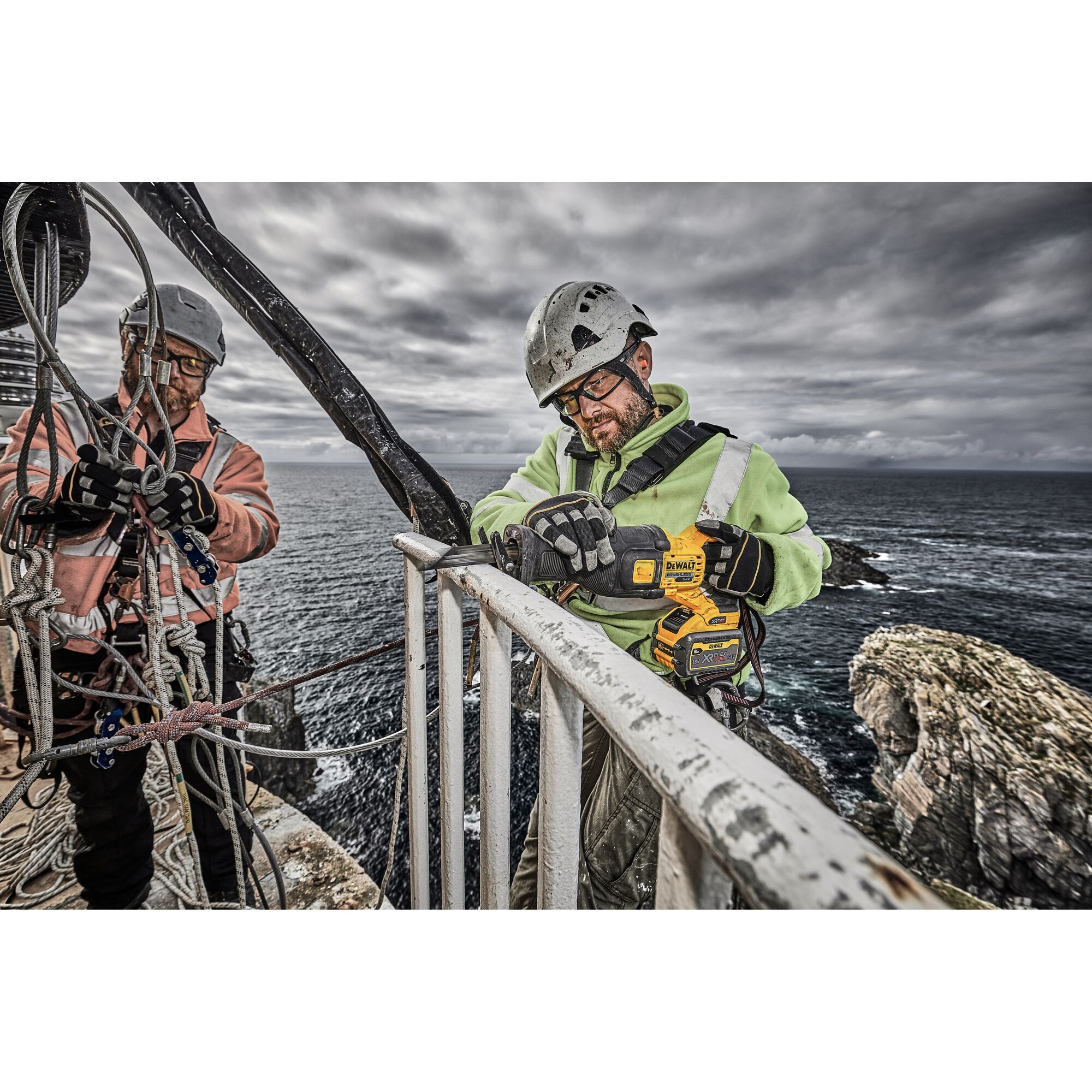 Two workers wearing safety helmets and harnesses are performing maintenance on a railing high above the sea, with one worker holding a DEWALT DCS389X2 power tool. The background shows a rugged coastline and cloudy sky.