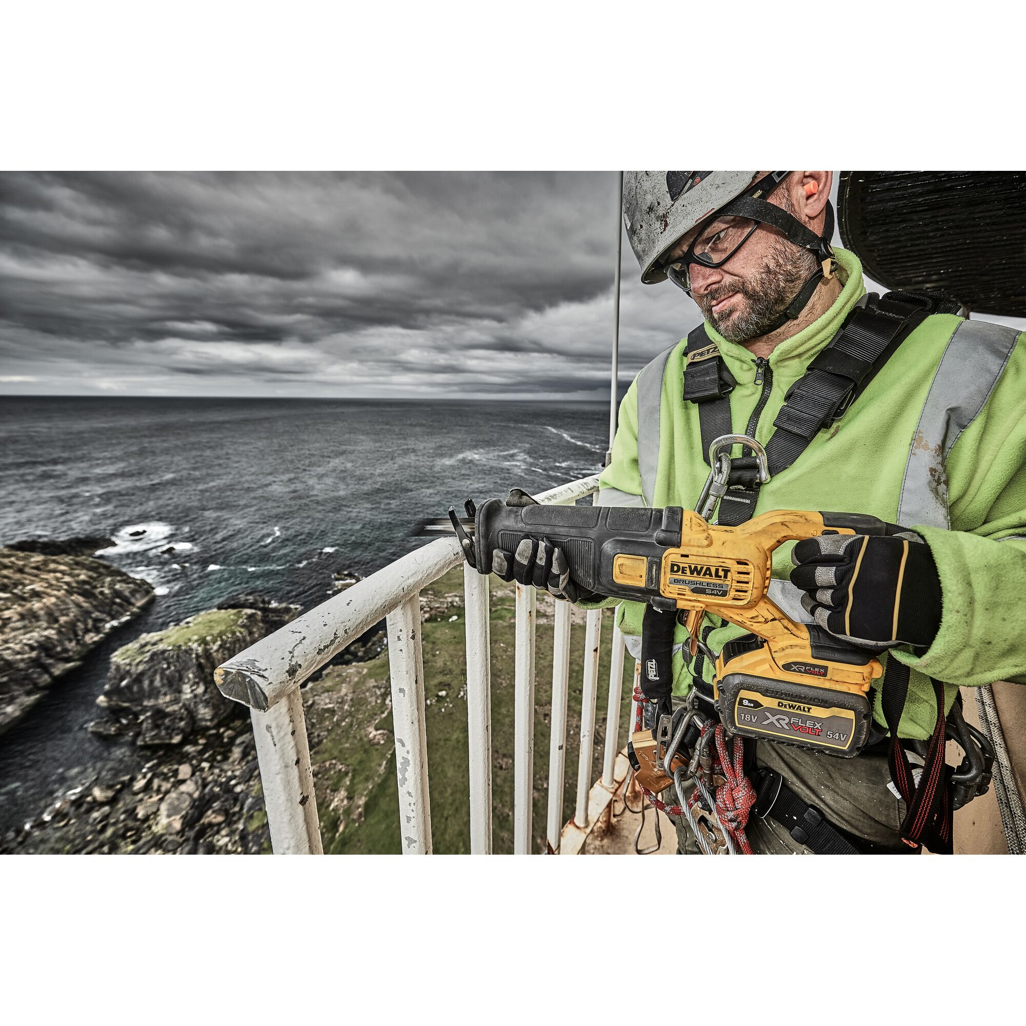 A worker in safety gear is holding a DEWALT cordless reciprocating saw (model DCS389X2) on an outdoor balcony overlooking the sea and rocky coastline. The worker's face is obscured.