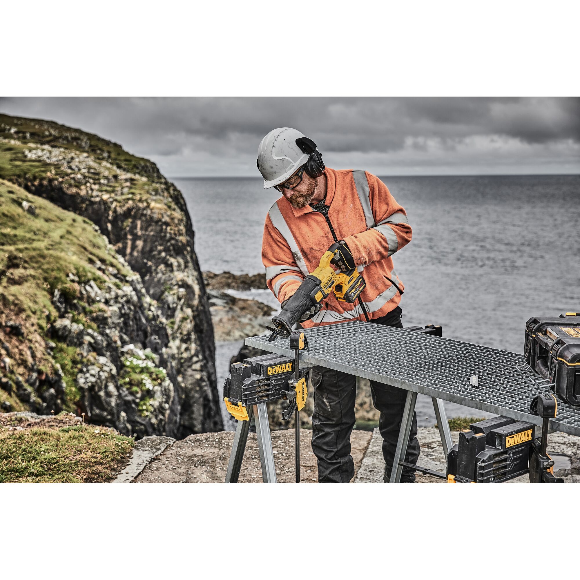 A worker wearing a helmet and an orange safety jacket is using a DEWALT cordless reciprocating saw on a metal workbench outdoors, near a rocky coastline.