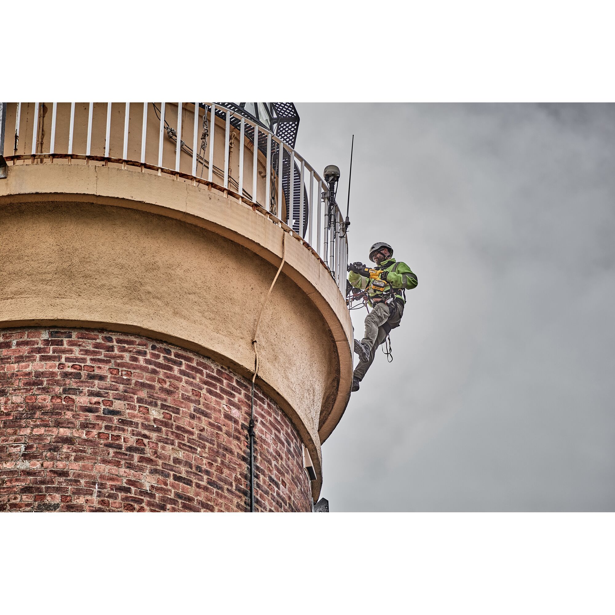 A worker equipped with safety gear is using a DEWALT power tool while suspended by ropes on the side of a brick and concrete structure, possibly performing maintenance or construction.