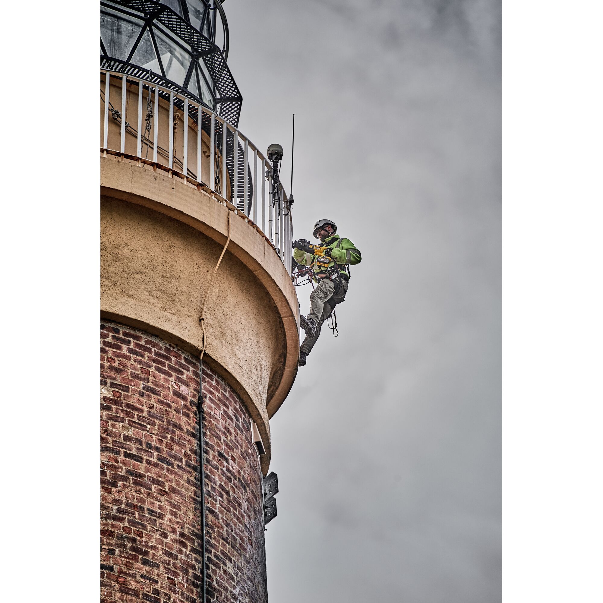 A person wearing safety gear is suspended on ropes, working on the exterior of a brick lighthouse with a railing near the top. The sky is cloudy.