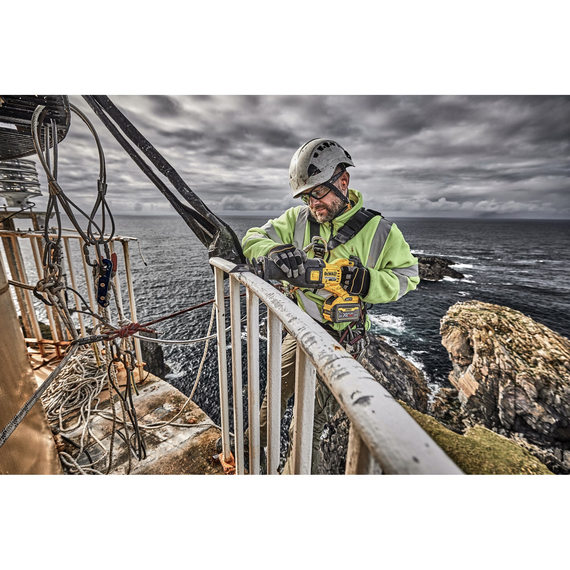 A person wearing safety gear is using a DEWALT DCS389X2 power tool to cut metal railing outdoors near the edge of a rocky coastline, with harnesses and ropes for safety.