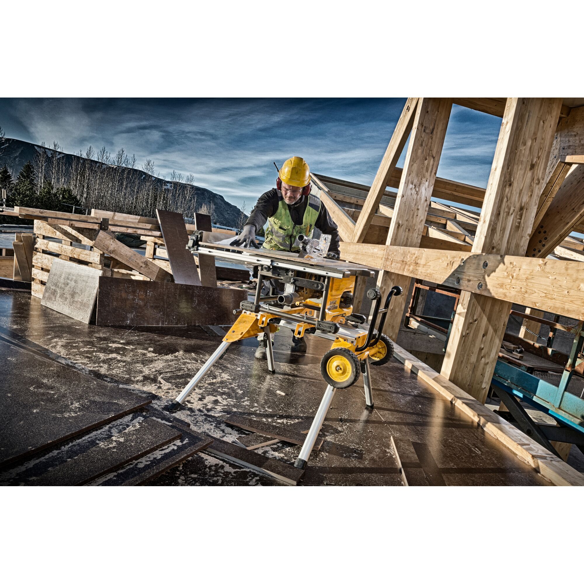 A construction worker using a Dewalt DCS7485N table saw on a jobsite, surrounded by wooden beams and structures, with protective gear and a mountain landscape in the background.