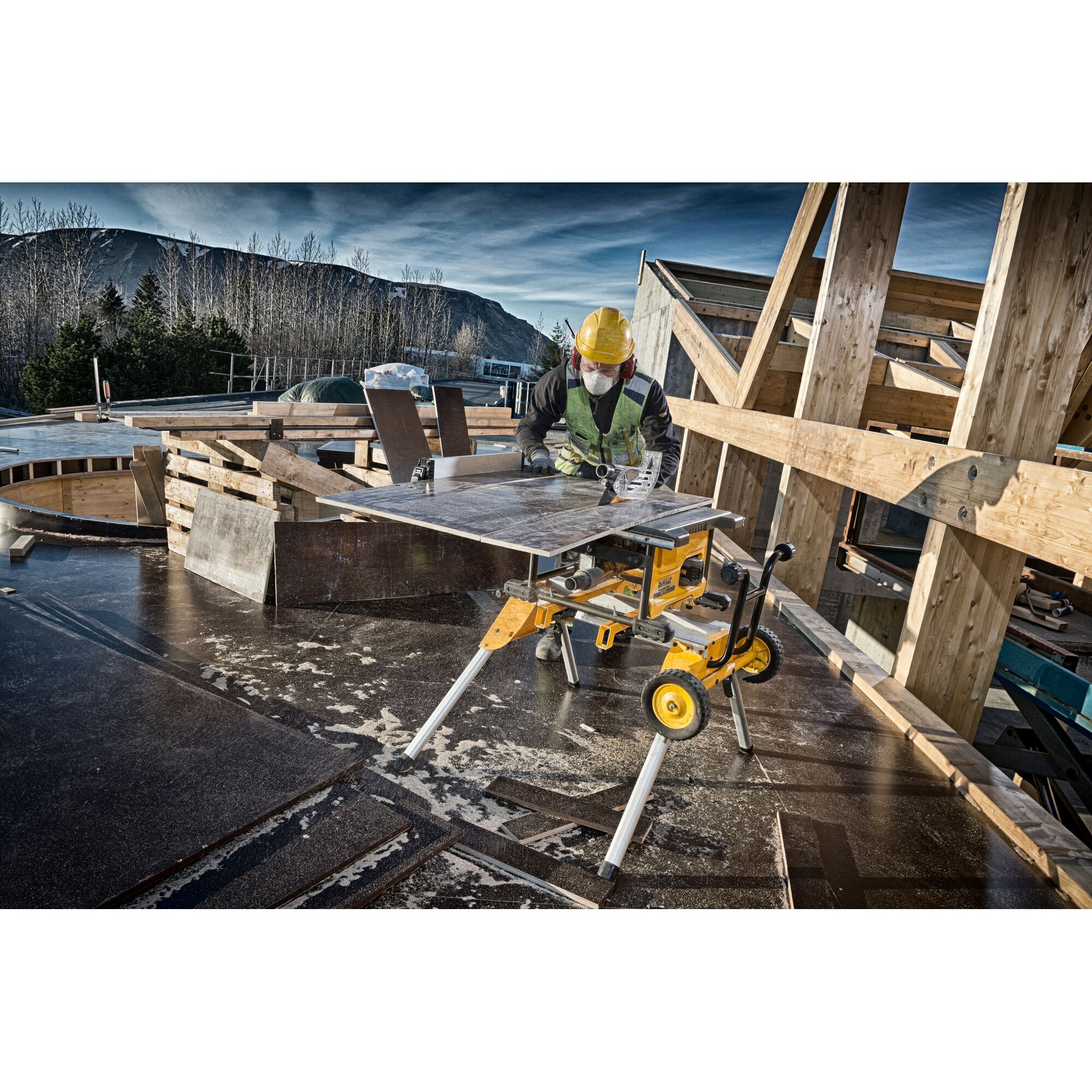 A worker in safety gear uses a DEWALT DCS7485N table saw outdoors at a construction site. Wood planks and construction materials surround the area, and a partially built wooden structure is visible in the background.