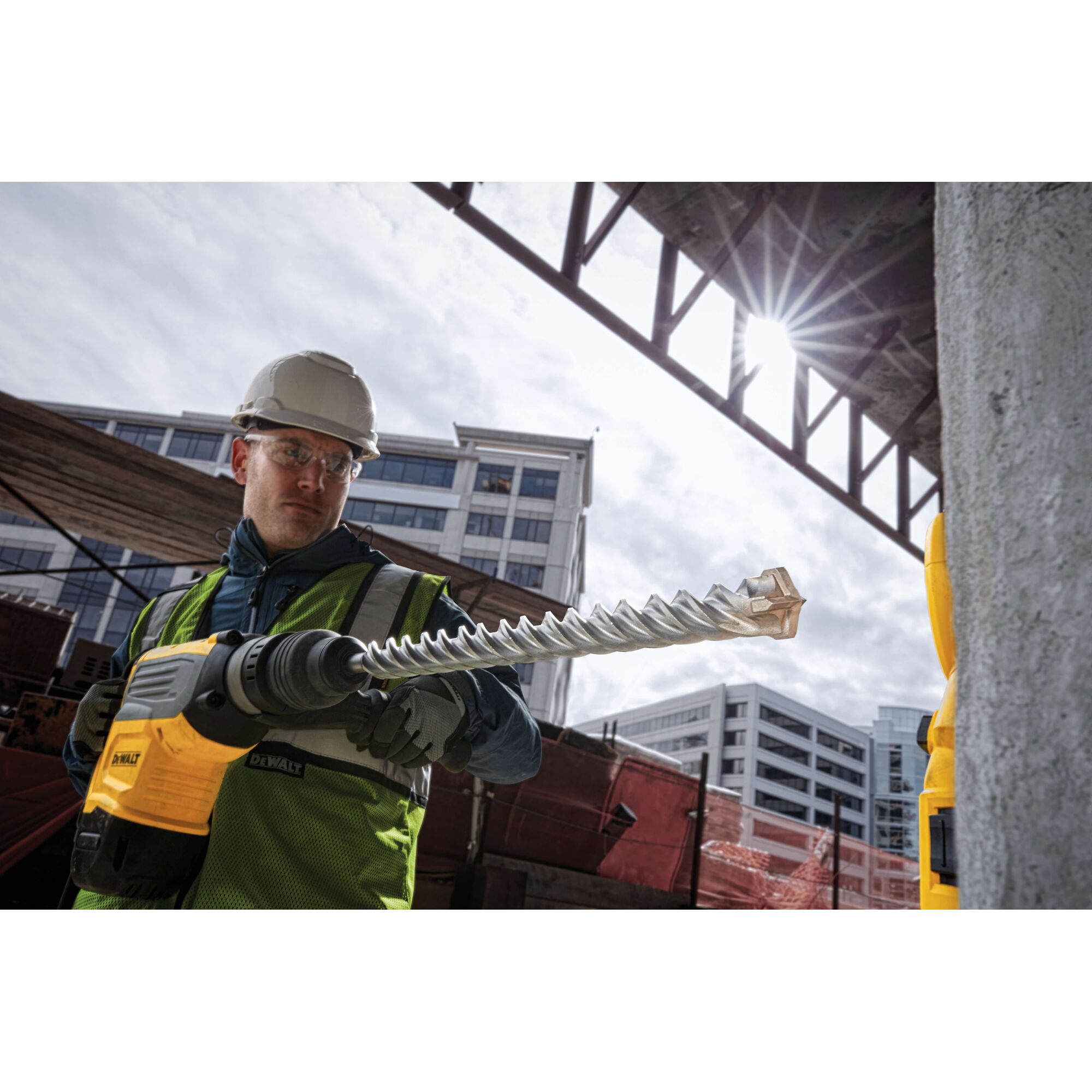 A construction worker wearing a safety helmet and high-visibility vest is holding a DEWALT power tool with a long, spiral drill bit on an outdoor building site. Modern office buildings are visible in the background, and the sun shines through a steel structure overhead.