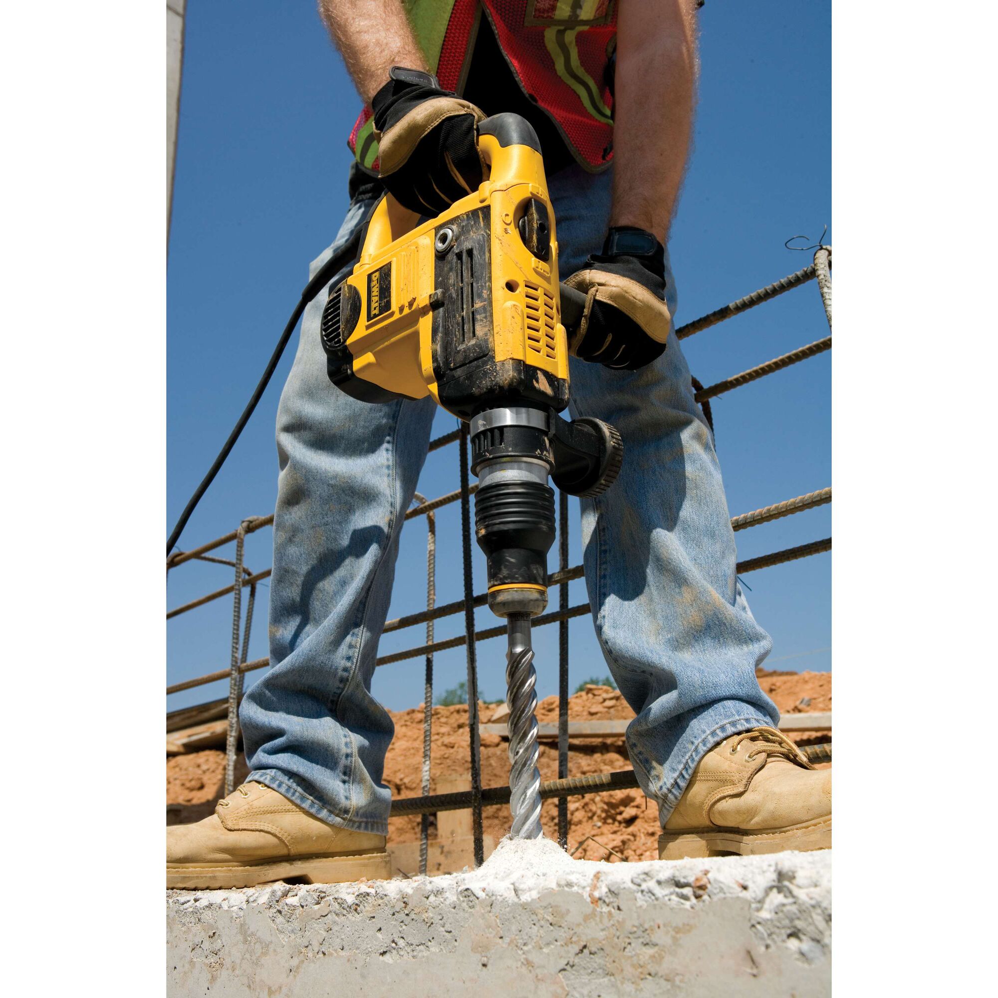 A person wearing gloves and construction attire uses a yellow DEWALT rotary hammer drill to drill into concrete on a construction site.