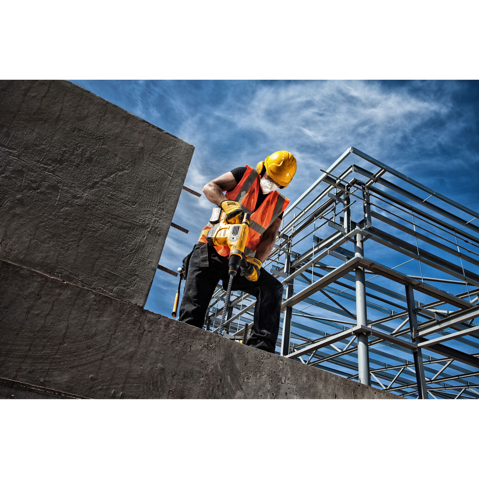 A construction worker wearing a yellow safety helmet, orange reflective vest, and face mask is operating a yellow DEWALT power tool, drilling into concrete at a construction site with steel framework and a blue sky in the background.