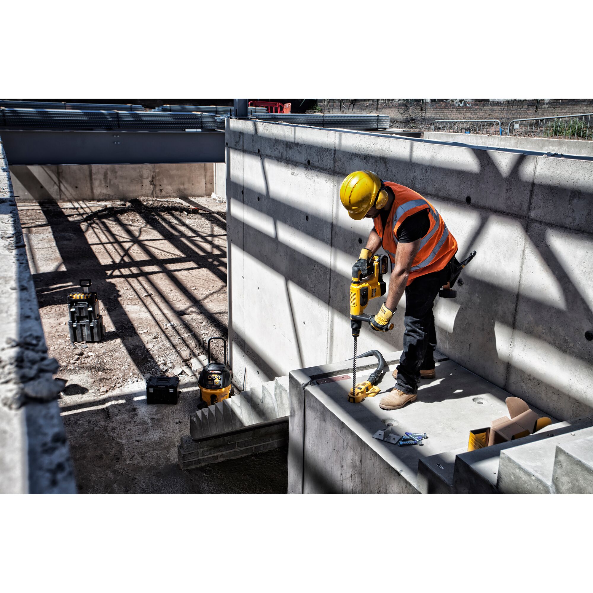 A construction worker wearing a yellow hard hat and an orange safety vest is using a DEWALT power tool to drill into a concrete structure at a construction site. Various tools and equipment are visible around the site.