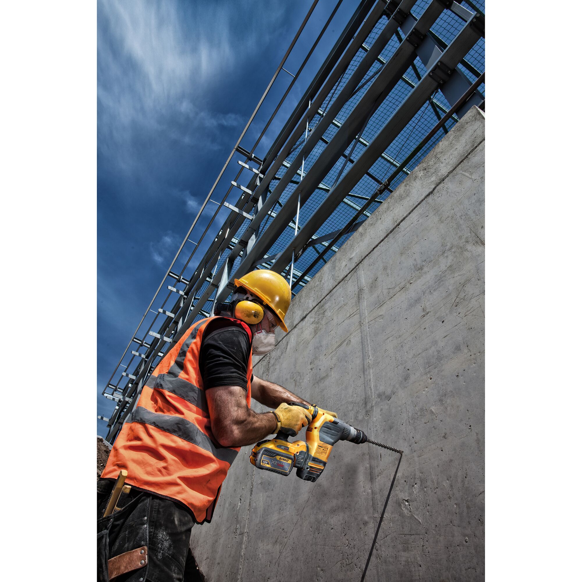A construction worker wearing a yellow hard hat, orange safety vest, and ear protection is using a DEWALT cordless rotary hammer drill to drill into a concrete wall outdoors, with blue sky and metal structures in the background.