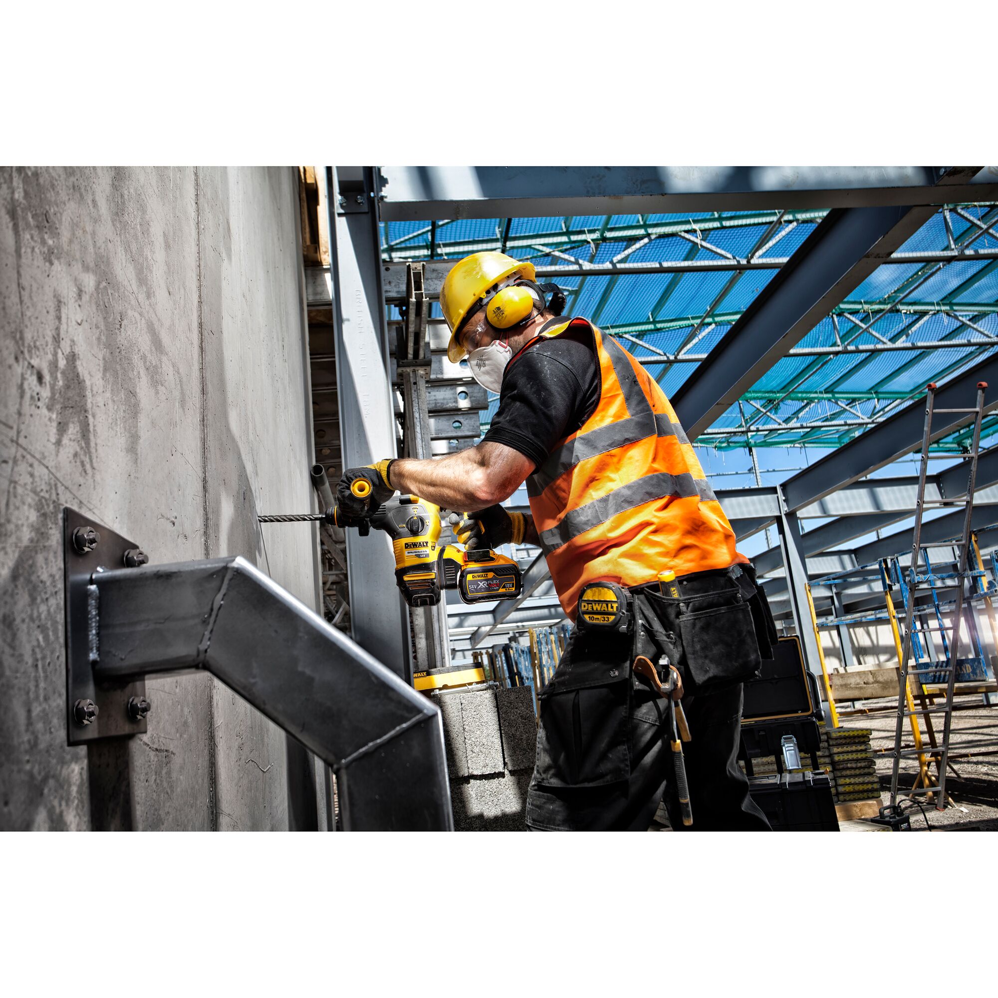 A construction worker wearing a yellow helmet, ear protection, and an orange safety vest is using a DEWALT power drill to drill into a concrete wall at an industrial construction site.