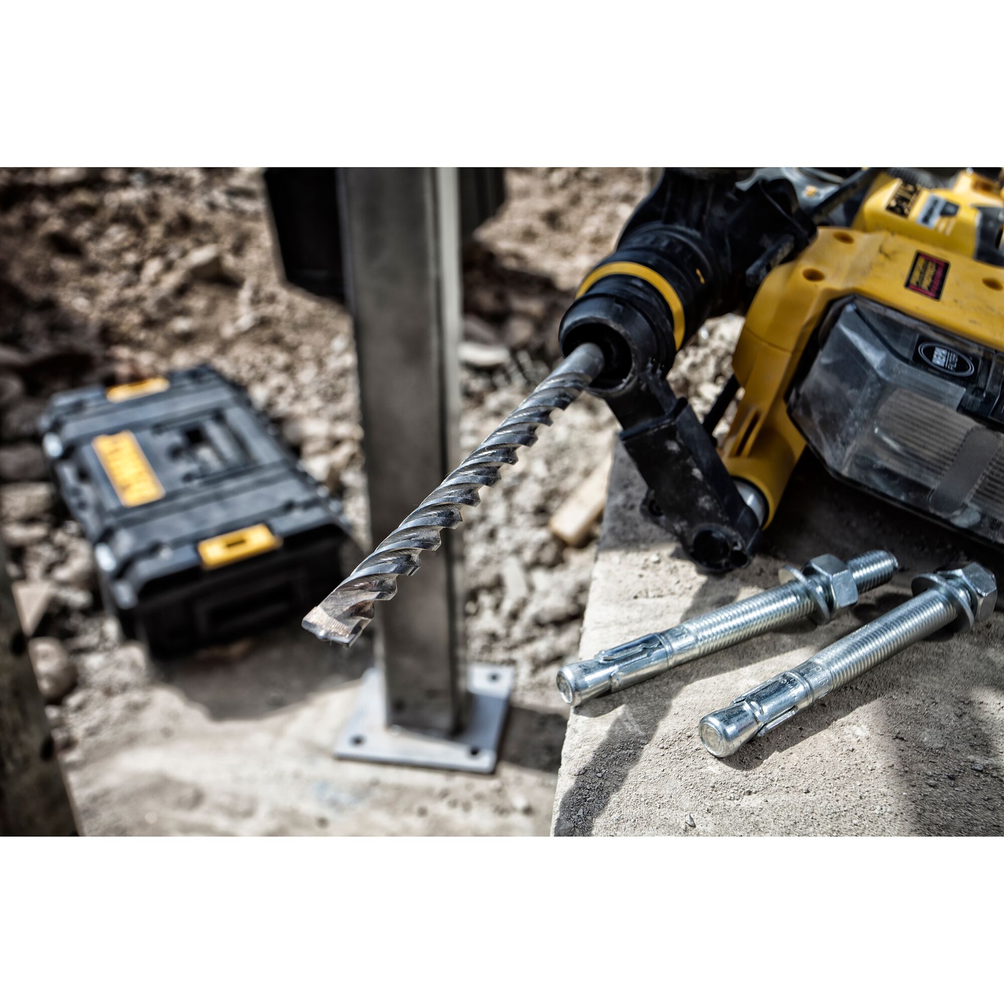 A close-up of a DEWALT power drill with a spiral masonry drill bit attached, lying on a concrete surface next to three large metal anchor bolts. In the background, a DEWALT branded toolbox sits on rocky ground at a construction site.