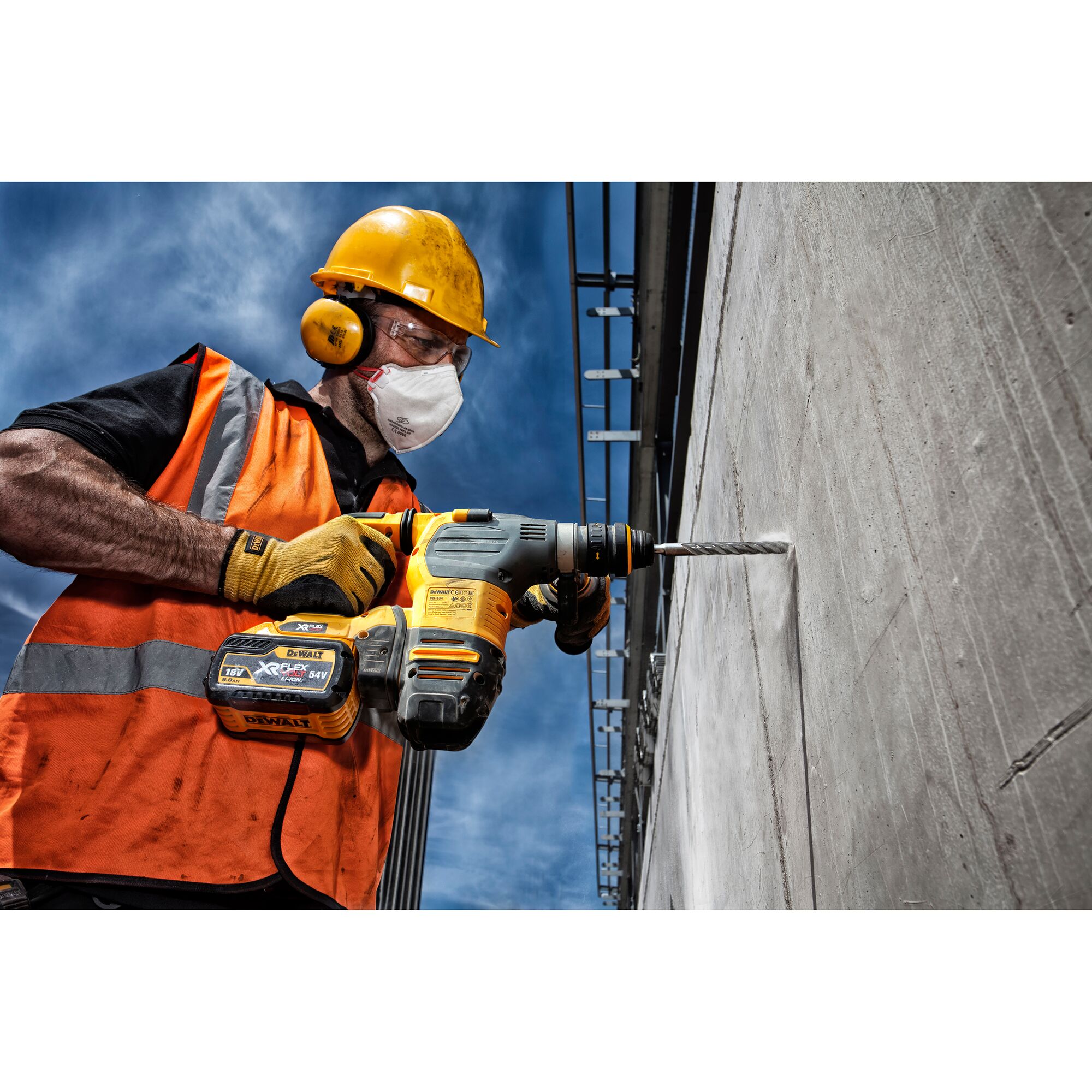 A construction worker wearing an orange safety vest, gloves, and a yellow hard hat uses a DEWALT cordless hammer drill to drill into a concrete wall outdoors under a blue sky.