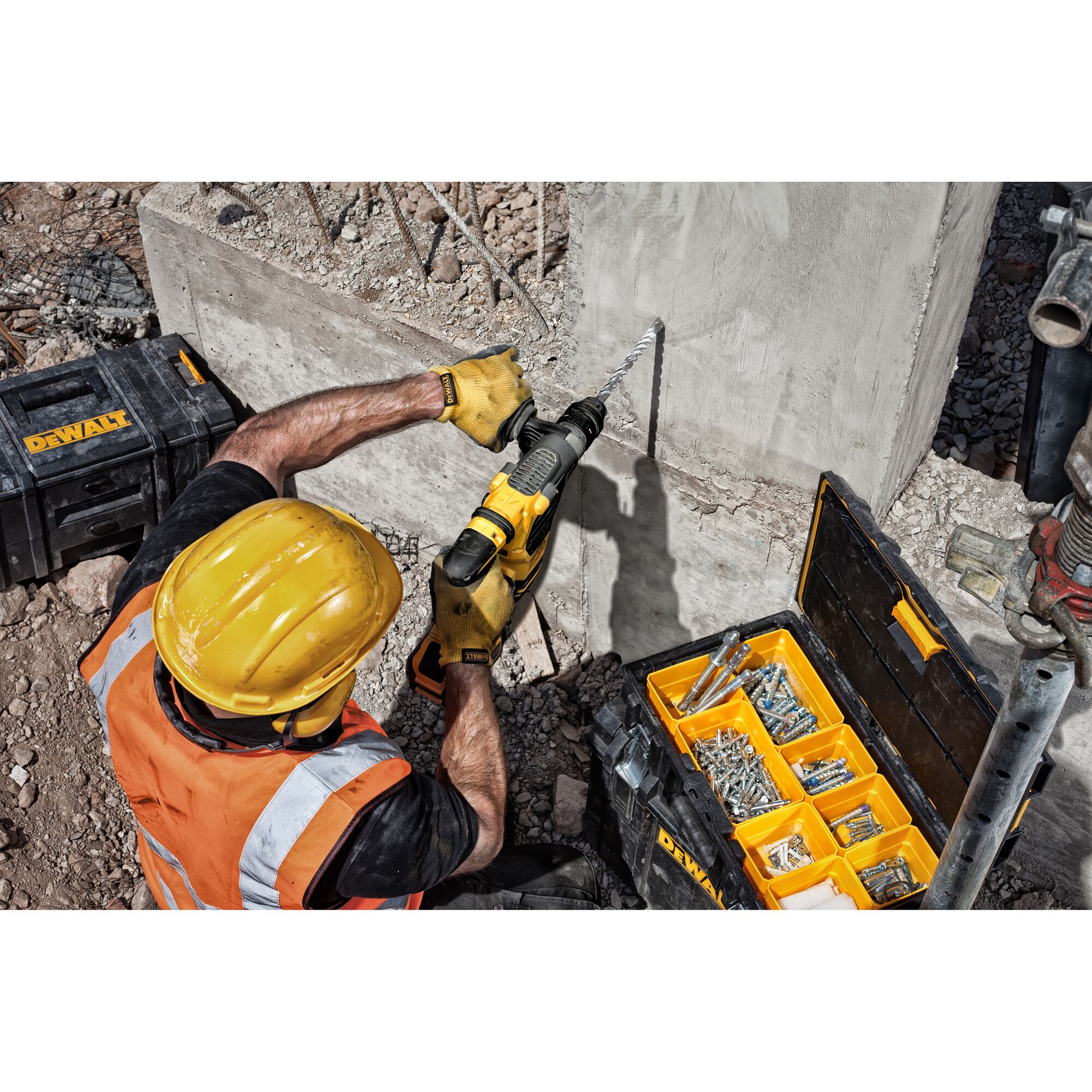 A construction worker wearing a yellow hard hat and orange safety vest uses a DEWALT power drill to drill into a concrete wall. There are toolboxes on the ground, one open containing various screws, bolts, and fixings.
