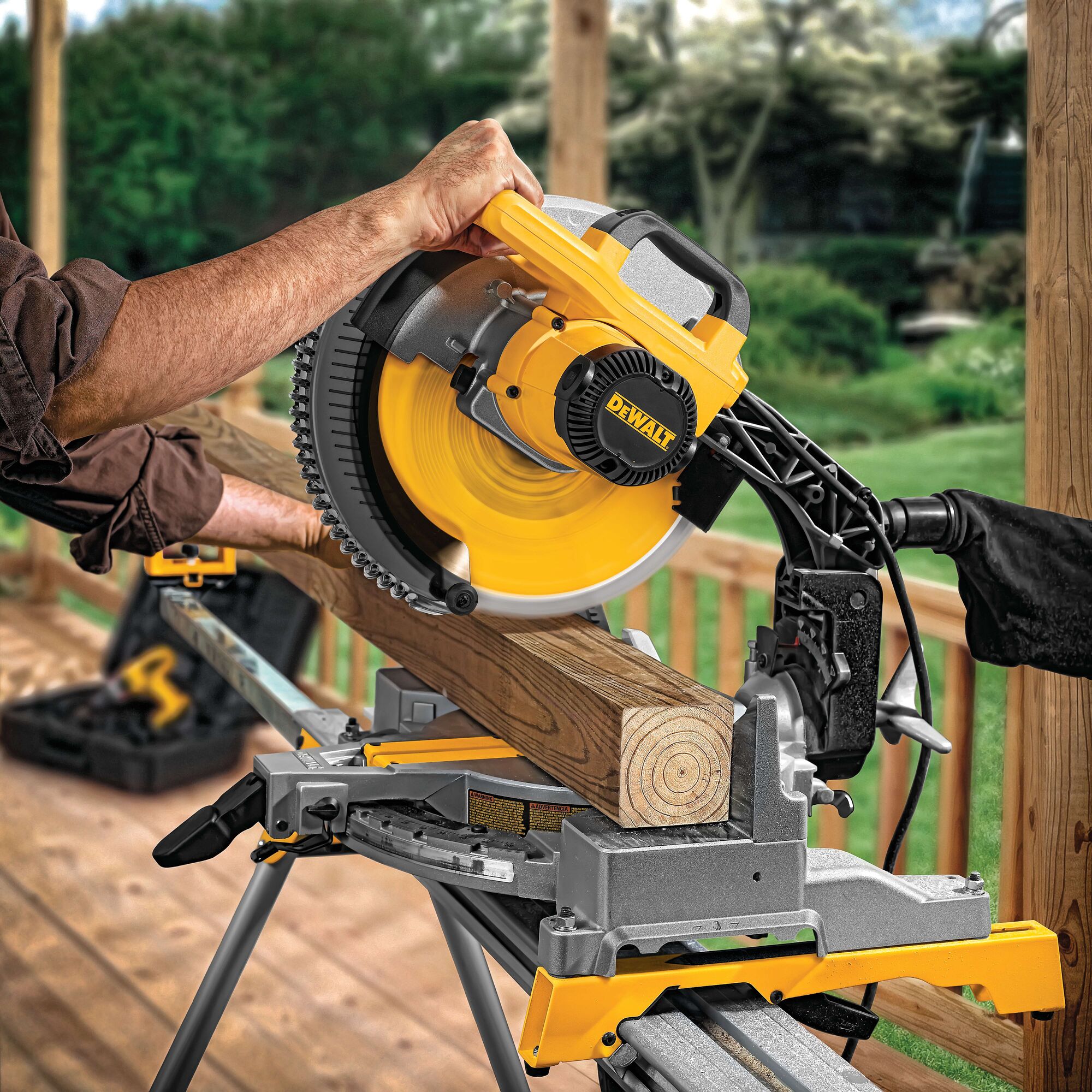 A person using a DEWALT miter saw to cut a piece of wood outdoors, with the saw mounted on a sturdy stand and surrounded by greenery.