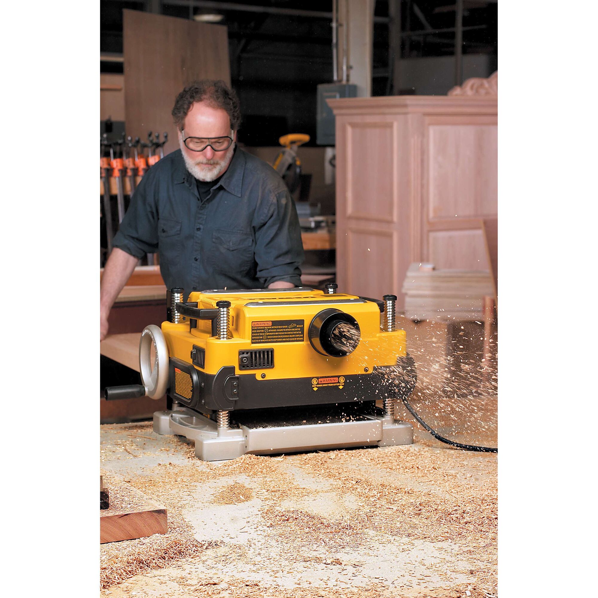 A DEWALT DW735 planer in use in a woodworking shop, creating wood shavings as it operates. A person stands behind the machine with their face blurred. The background shows wooden cabinets and various tools.